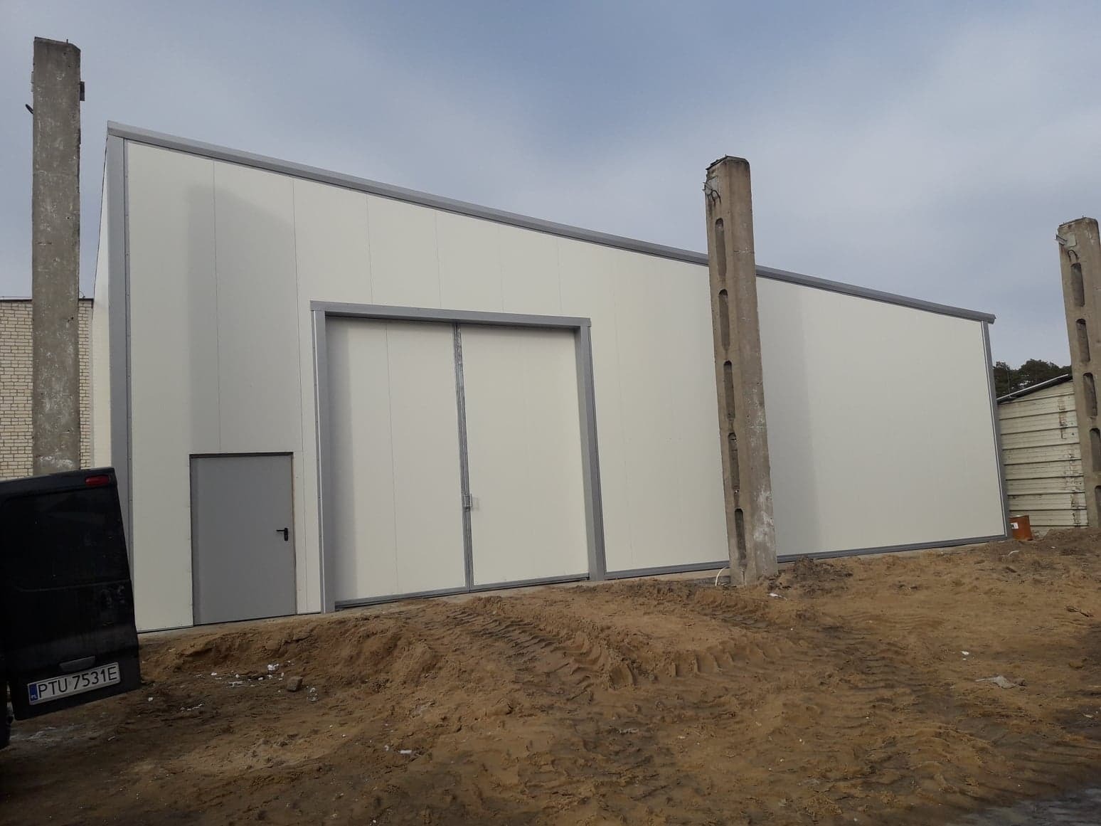 Nowo wybudowana hala z płyt warstwowych z widocznymi drzwiami przesuwnymi i wejściowymi, na placu budowy z widocznym samochodem dostawczym i słupami betonowymi.