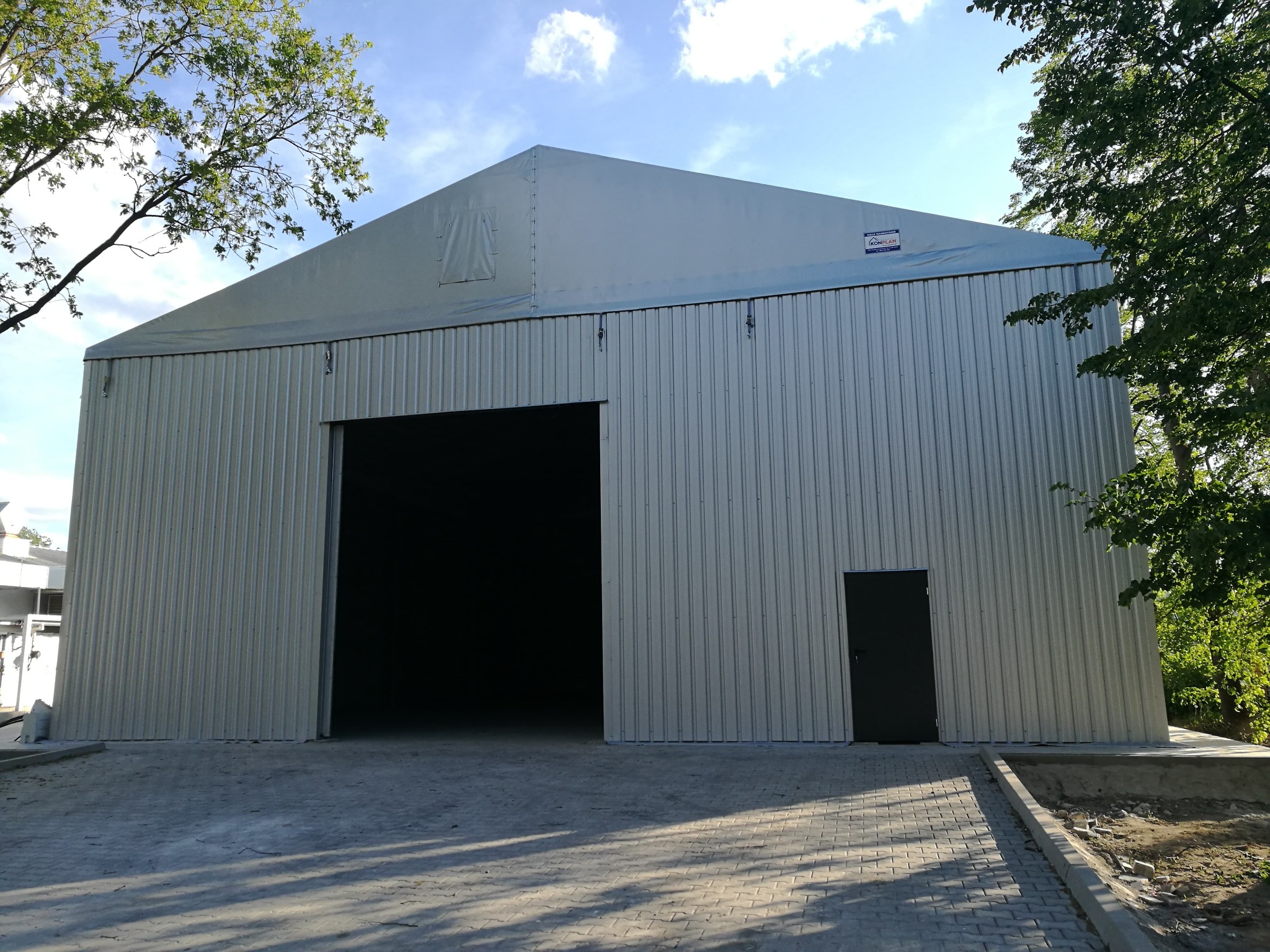 Gotowa hala stalowa z blachy trapezowej, z dużym otwartym wjazdem i pojedynczymi drzwiami, widoczna tabliczka informacyjna na górze, otoczenie zieleni i kostka brukowa.