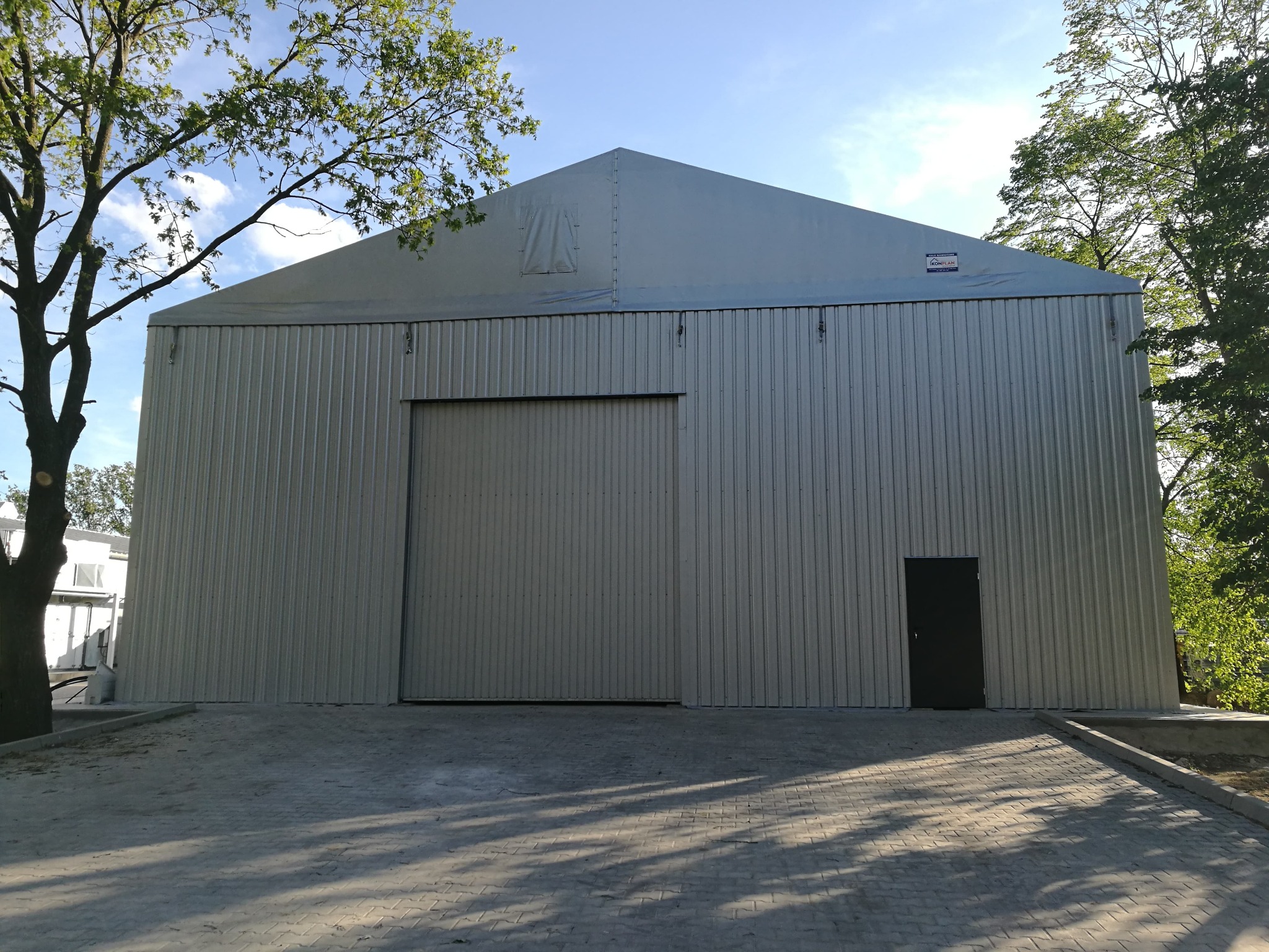 Gotowa hala magazynowa z szarej blachy trapezowej, z dużymi przesuwnymi drzwiami i dodatkowym wejściem, widoczna tabliczka informacyjna na górze ściany szczytowej.