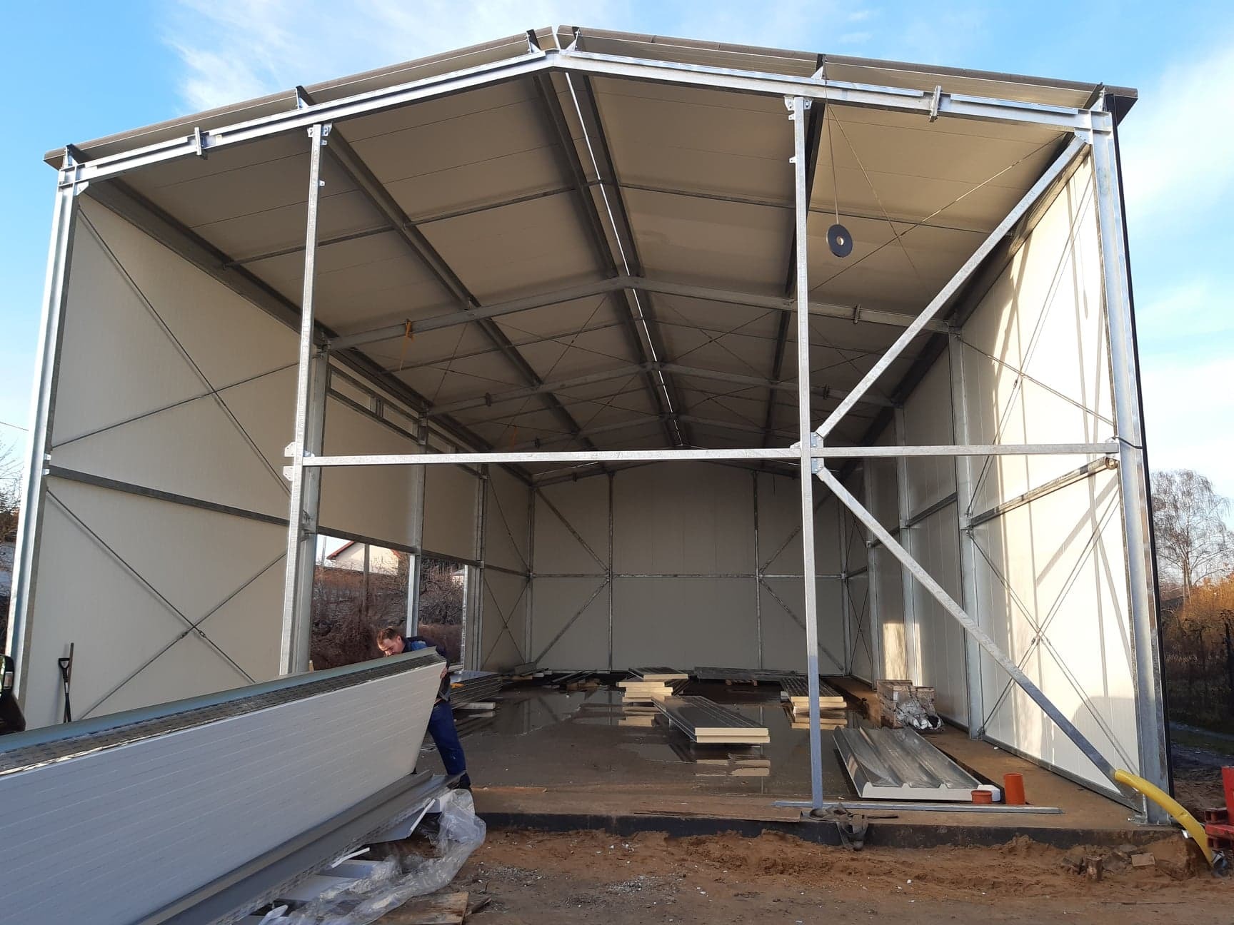 Konstrukcja hali w trakcie budowy z widocznym pracownikiem przenoszącym płytę warstwową. Wnętrze hali z widocznymi elementami konstrukcyjnymi i materiałami budowlanymi.