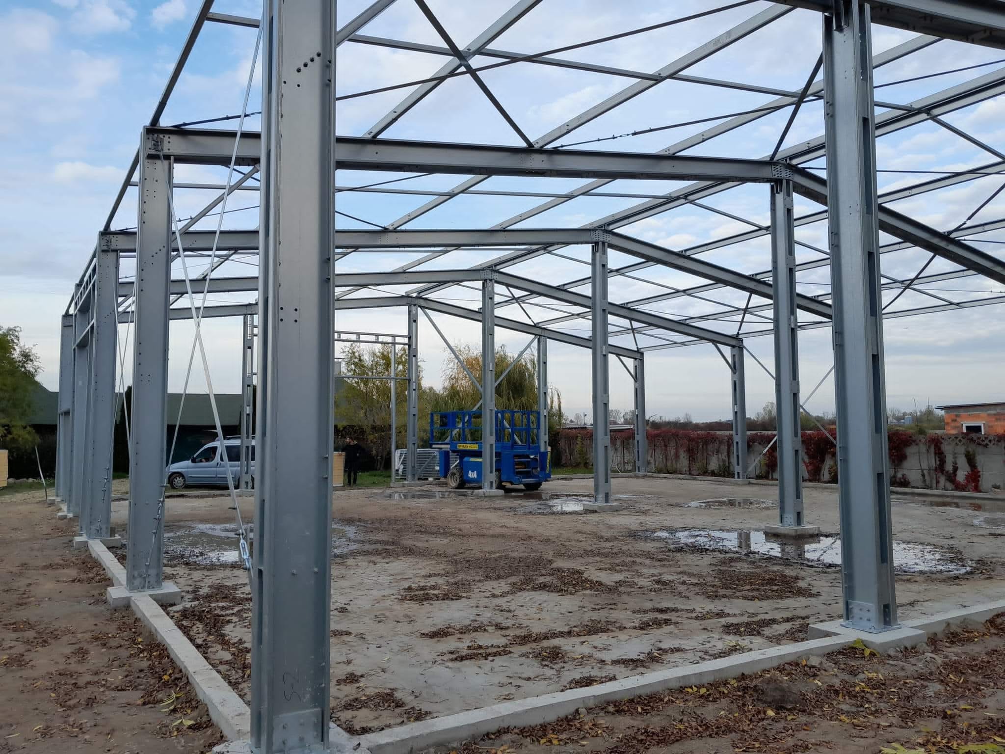 Konstrukcja stalowa hali w trakcie budowy, widoczne słupy i kratownica, w tle podnośnik koszowy i częściowo zabudowany teren.
