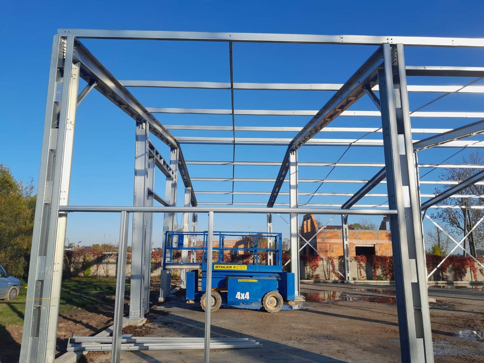 Stalowa konstrukcja hali w trakcie budowy, widoczny podnośnik koszowy 4x4 na placu budowy, w tle częściowo zrujnowany budynek z czerwonej cegły.