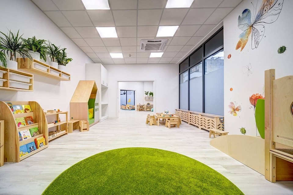 Jasne, przestronne wnętrze placówki edukacyjnej z drewnianymi meblami, zielonym dywanem w kształcie półkola i ścianą ozdobioną motywem motyli.