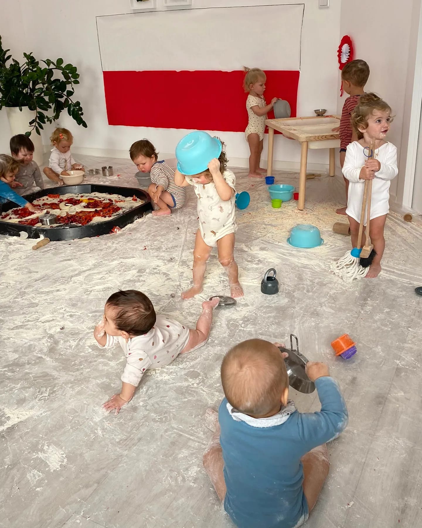 Grupa małych dzieci w jasnych ubraniach bawi się w pomieszczeniu z białą podłogą pokrytą mąką, jedno dziecko ma na głowie niebieską miskę, w tle widoczna flaga Polski i duża taca z owocami.