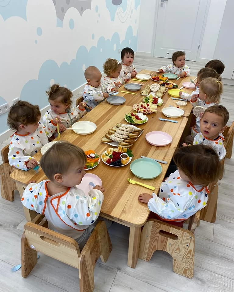 Grupa dzieci w jednakowych śliniakach siedzi przy drewnianym stole z posiłkiem, widok z góry.