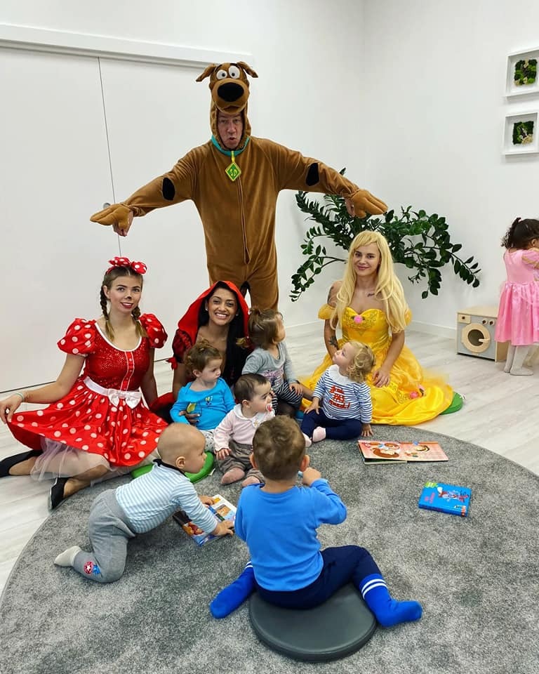 Animatorki przebrane za postacie z bajek, Scooby Doo, Królewna Śnieżka i Czerwony Kapturek, bawiące się z grupą małych dzieci na szarym dywanie w żłobku.