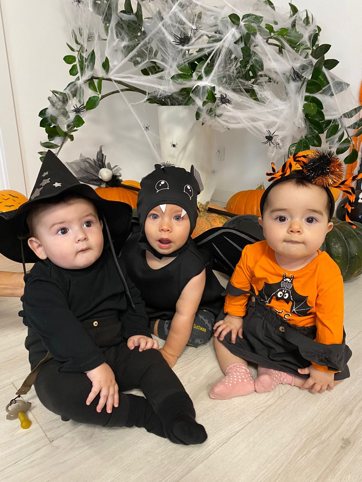Trzy małe dzieci w kostiumach na Halloween: czarownica z smoczkiem, nietoperz i dziewczynka z pająkiem na opasce, siedzą przed dyniami i dekoracjami z pajęczyn.