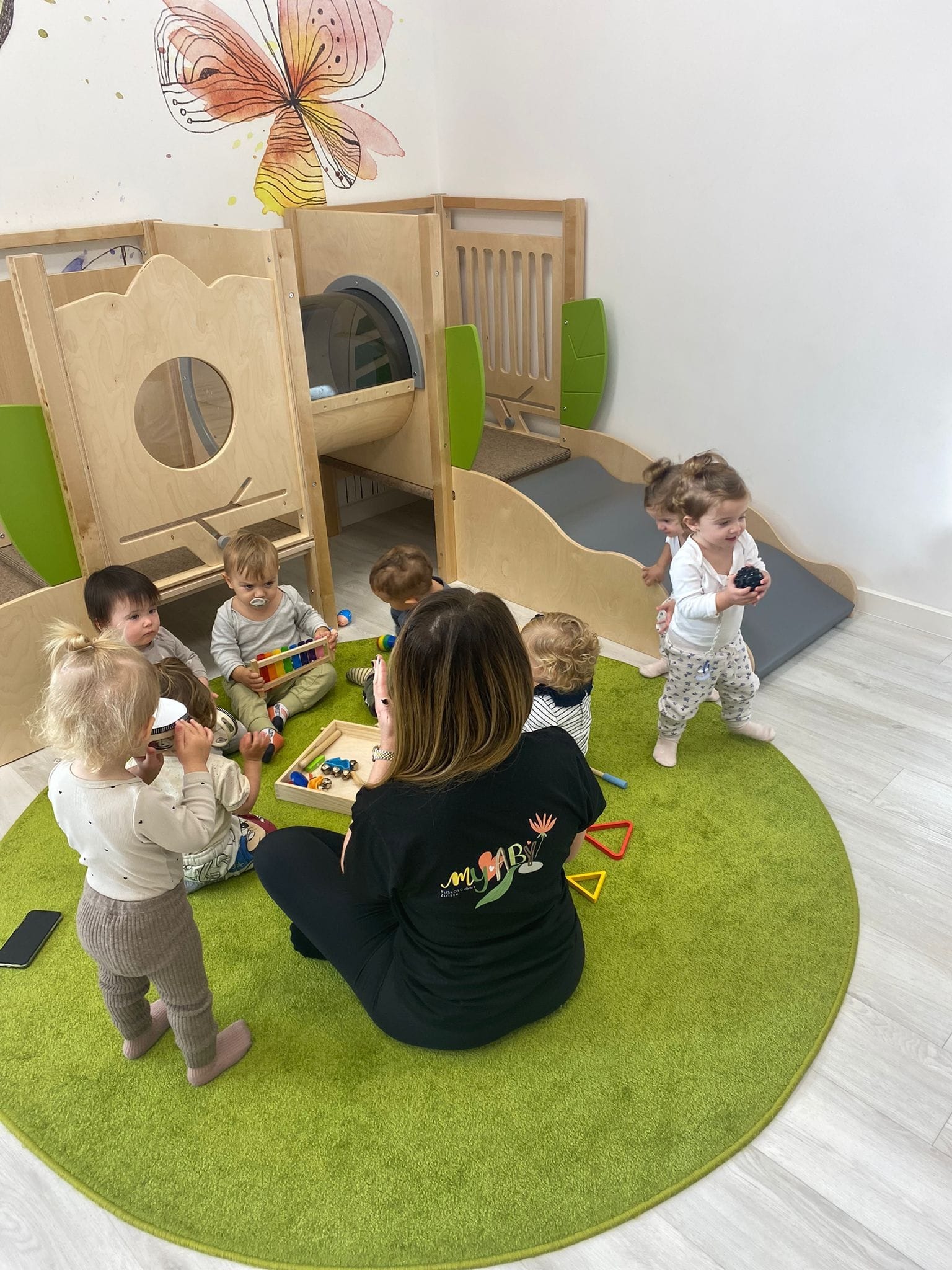 Grupa dzieci w jasnym pokoju zabaw z drewnianą konstrukcją do wspinania i zjeżdżalnią, siedzących na zielonym dywanie z opiekunką, która ma na sobie czarną koszulkę z kolorowym nadrukiem.