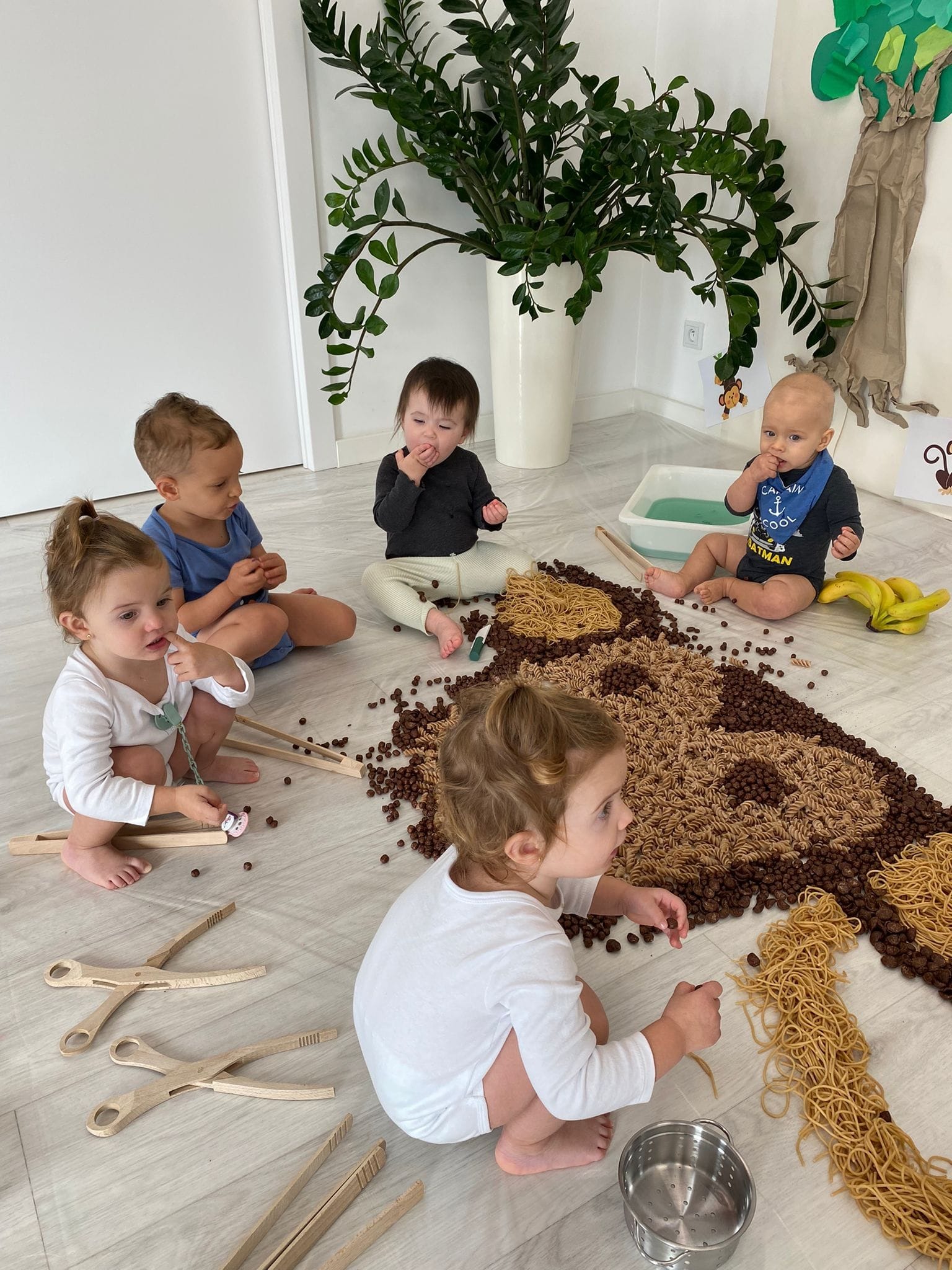 Pięcioro małych dzieci siedzi na podłodze w jasnym pokoju, bawiąc się makaronem spaghetti, brązowymi kulkami zbożowymi i drewnianymi szczypcami, tworząc kompozycję przypominającą niedźwiedzia...