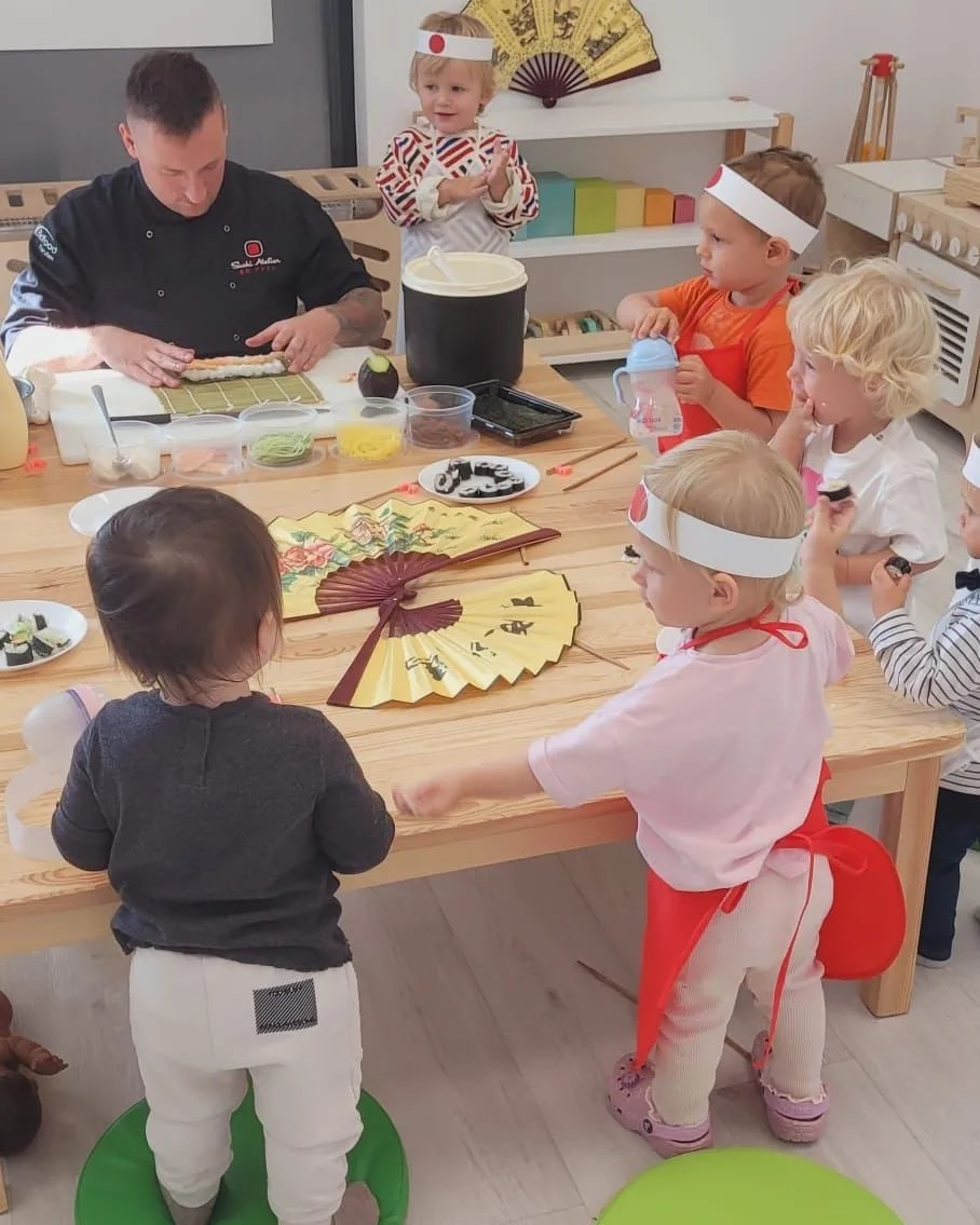 Dzieci w opaskach z motywem japońskiej flagi obserwują szefa kuchni przygotowującego sushi na drewnianym stole, z dekoracyjnymi wachlarzami i kolorowymi pojemnikami z jedzeniem.