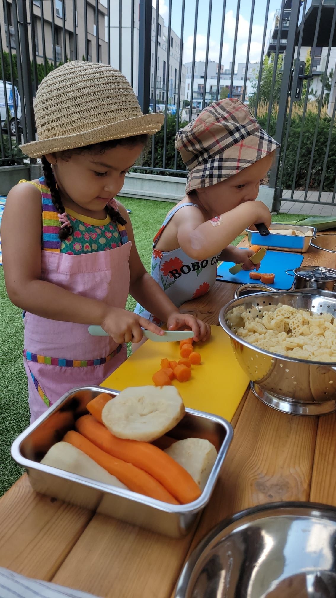 Dwoje dzieci w kapeluszach kroi warzywa na żółtej desce podczas zajęć kulinarnych na świeżym powietrzu; w tle budynki mieszkalne.
