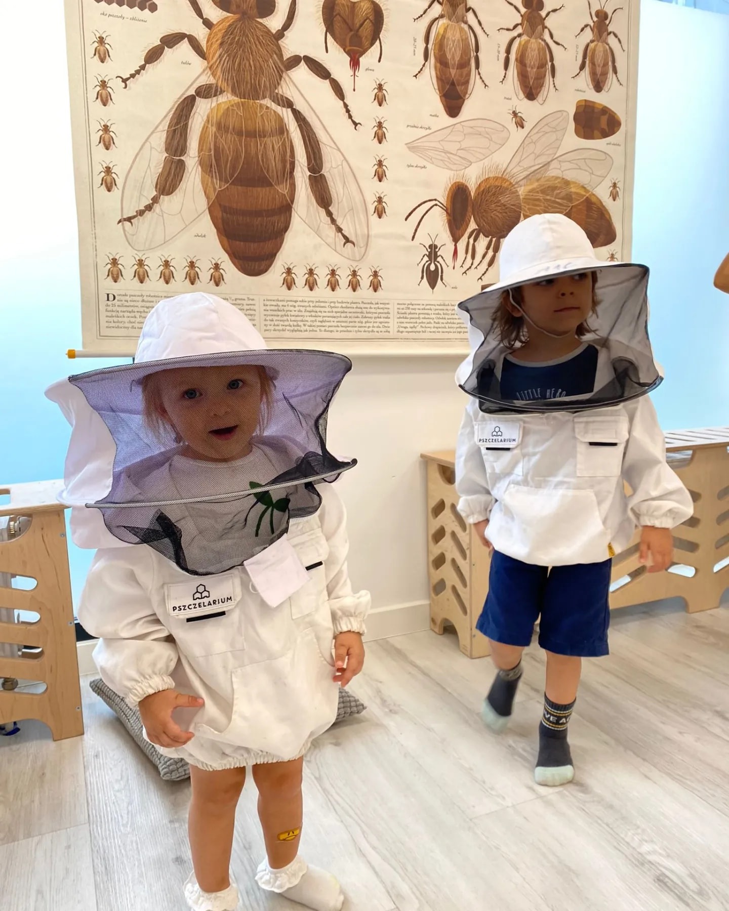 Dwoje dzieci ubranych w białe kombinezony pszczelarskie z siatkowymi kapeluszami, stojących na tle plakatu z ilustracjami pszczół.