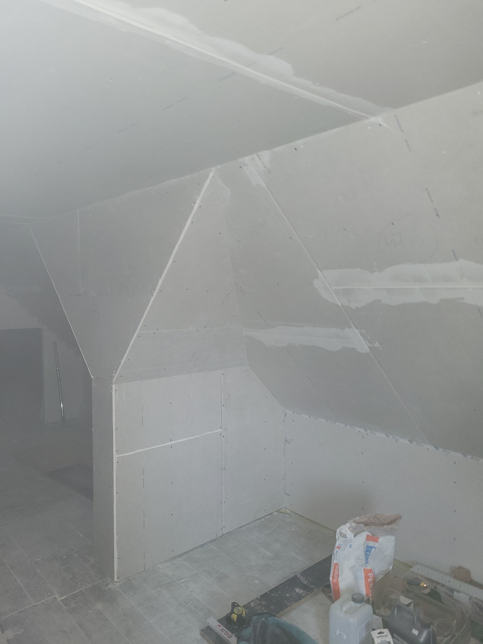 Montaż płyt karton-gips na poddaszu ze skosami. Widoczne łączenia płyt i narzędzia pracy. W tle widoczne wejście do innego pomieszczenia.