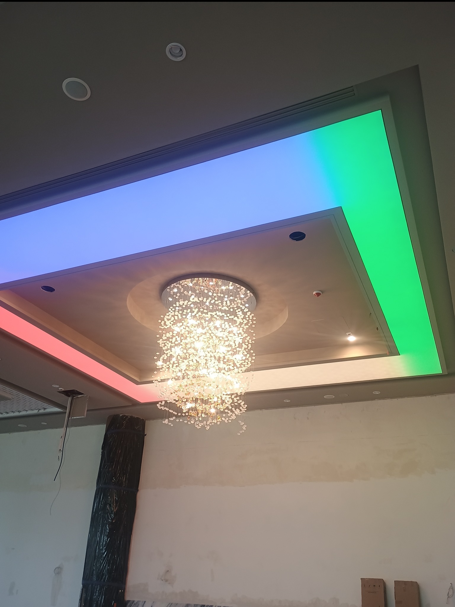 Podwieszany sufit z oświetleniem LED RGB w kolorach czerwonym, niebieskim i zielonym, z centralnie umieszczonym, kryształowym żyrandolem. Widoczne elementy wykończeniowe.