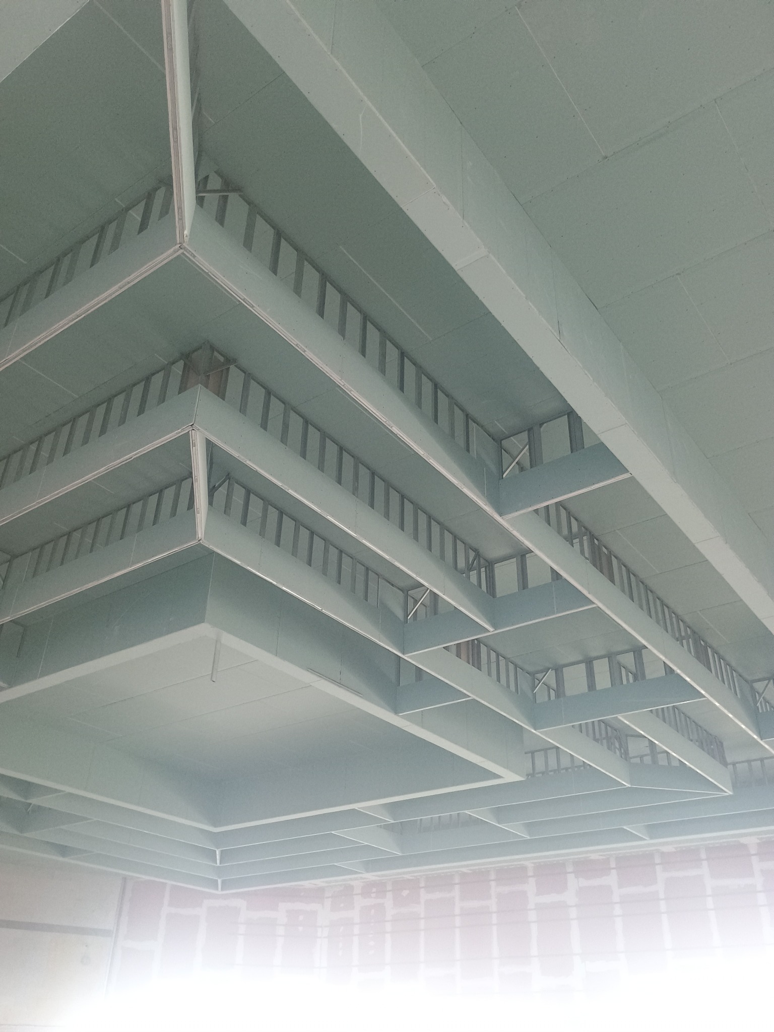 Skomplikowana konstrukcja sufitu podwieszanego z płyt gipsowo-kartonowych, widok z dołu, ukazujący metalowy stelaż i fragment ściany w tle.