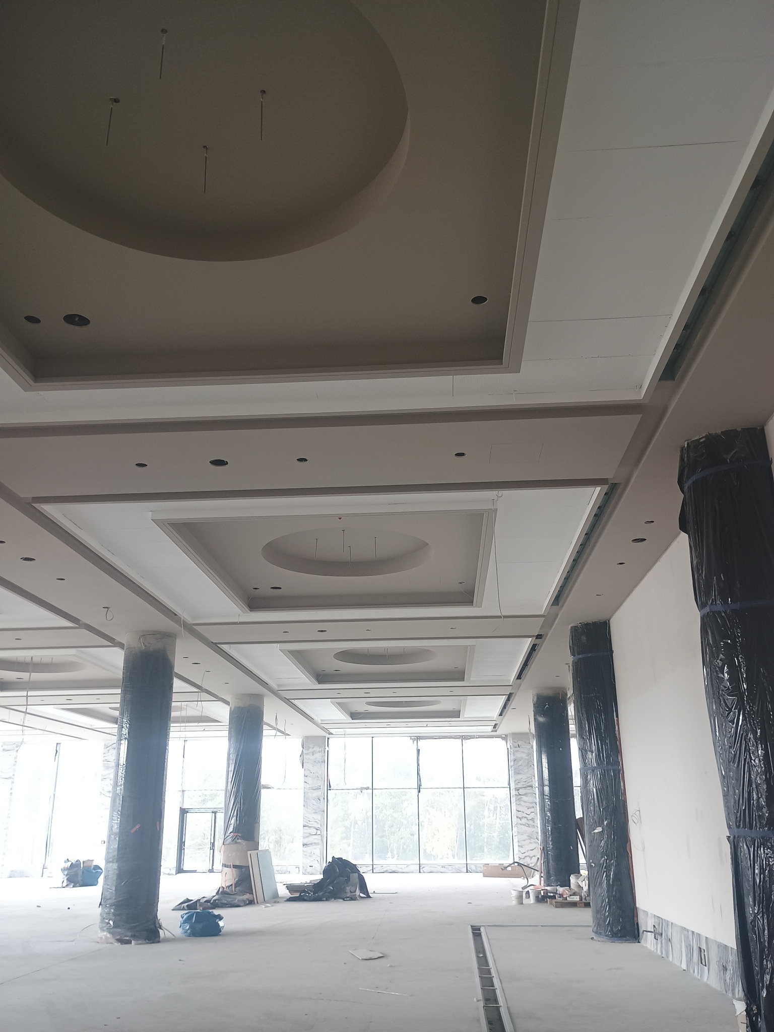 Wnętrze w trakcie wykończenia: dekoracyjny sufit z elementami kartonowo-gipsowymi i kolumny zabezpieczone folią. Duże okna w tle, surowy stan podłogi.