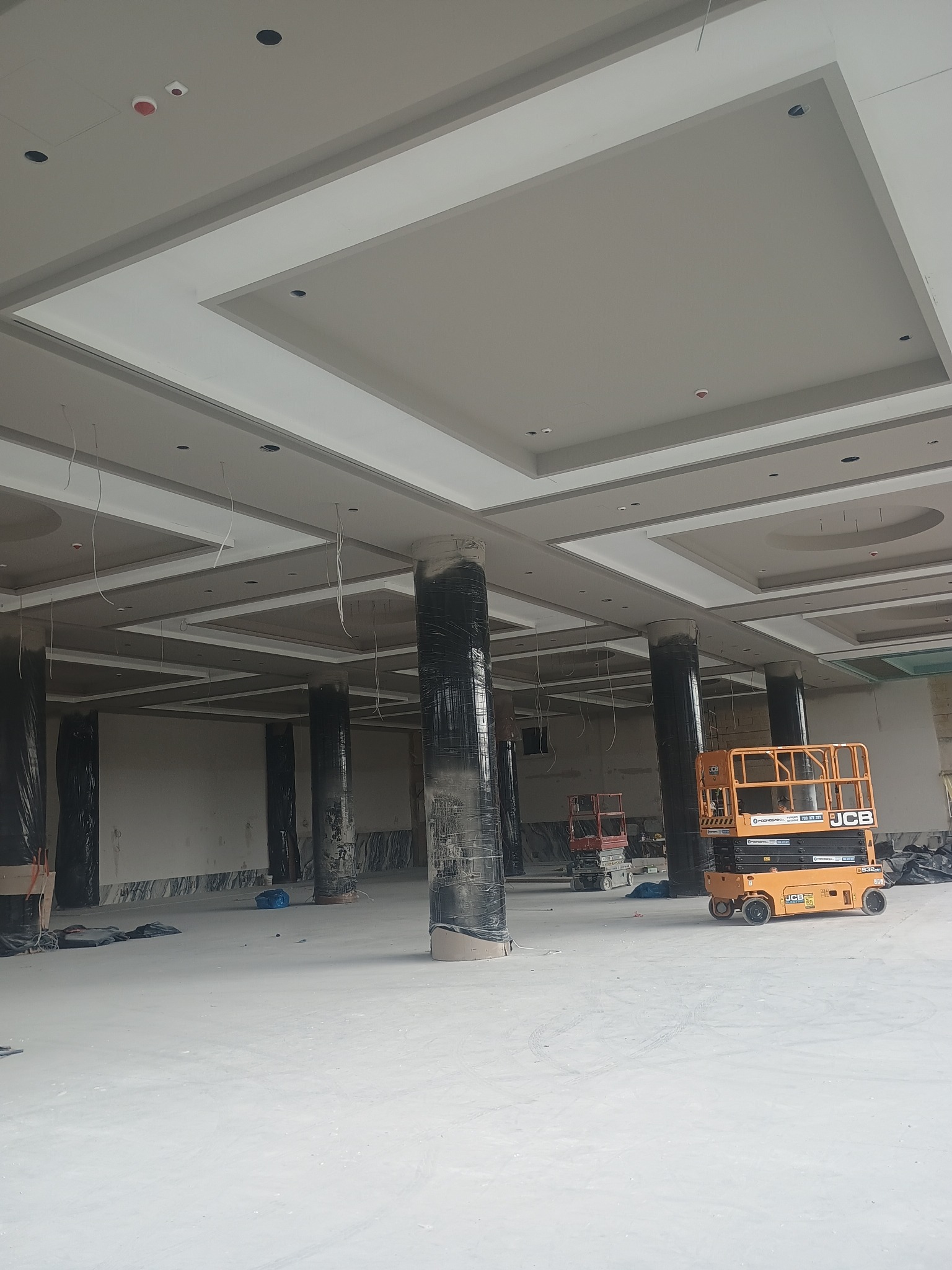 Montaż sufitu kasetonowego w dużej przestrzeni. Widoczne kolumny zabezpieczone folią, podnośniki i instalacje elektryczne. Prace wykończeniowe w toku.