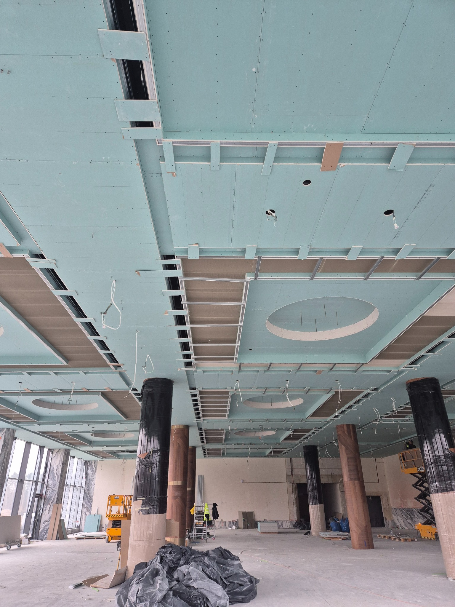 Montaż sufitu podwieszanego z płyt gipsowo-kartonowych w przestronnym wnętrzu. Widoczne metalowe profile i otwory na oświetlenie. W tle kolumny i podnośnik.