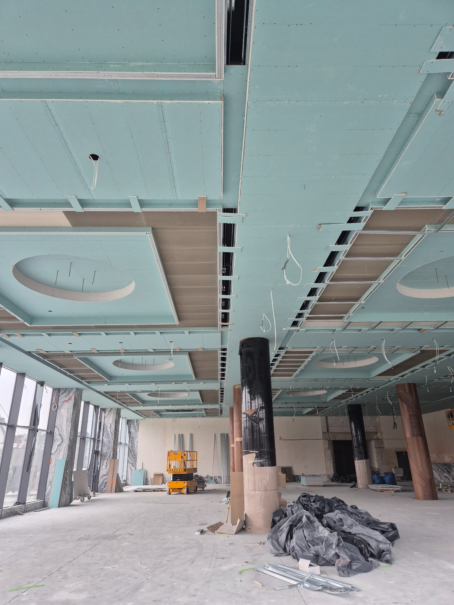 Montaż sufitu podwieszanego z płyt gipsowo-kartonowych z okrągłymi elementami dekoracyjnymi. Widoczna konstrukcja nośna i częściowo zamontowane płyty. W tle podnośnik.