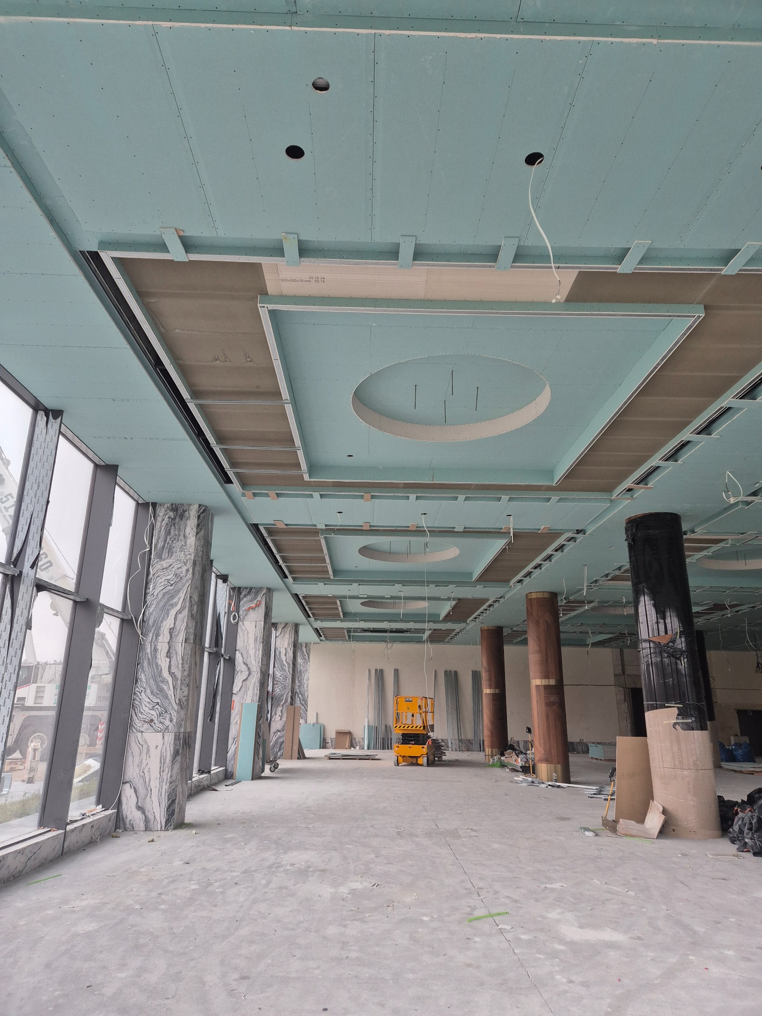 Montaż sufitu podwieszanego z płyty GK w przestronnym wnętrzu. Widoczne okrągłe elementy dekoracyjne i podkonstrukcja. W tle podnośnik i kolumny.