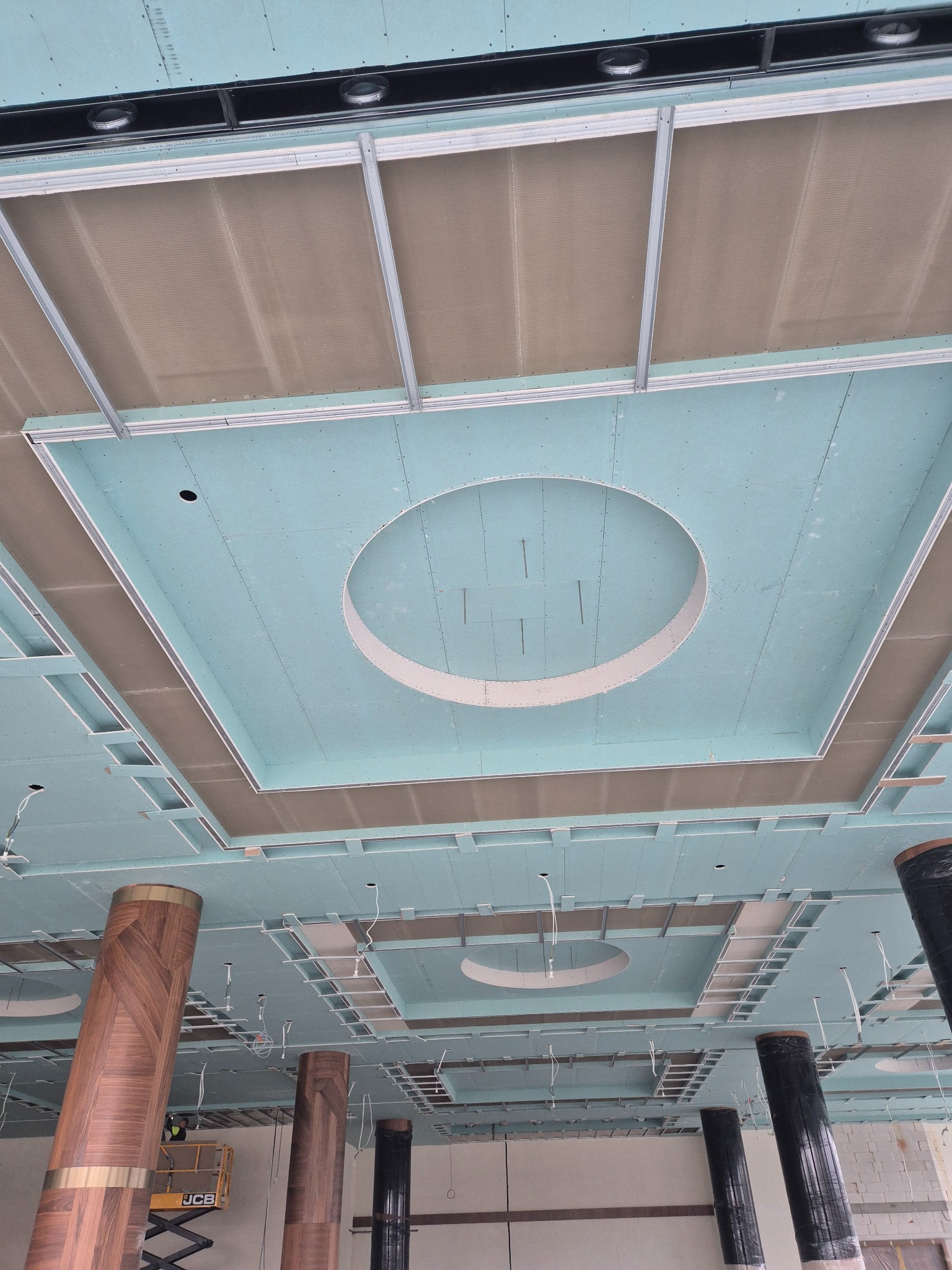 Montaż sufitu podwieszanego z płyty gipsowej z okrągłymi wnękami na oświetlenie. Widoczne metalowe profile i kolumny w dekoracyjnej okleinie. Prace wykończeniowe w obiekcie.