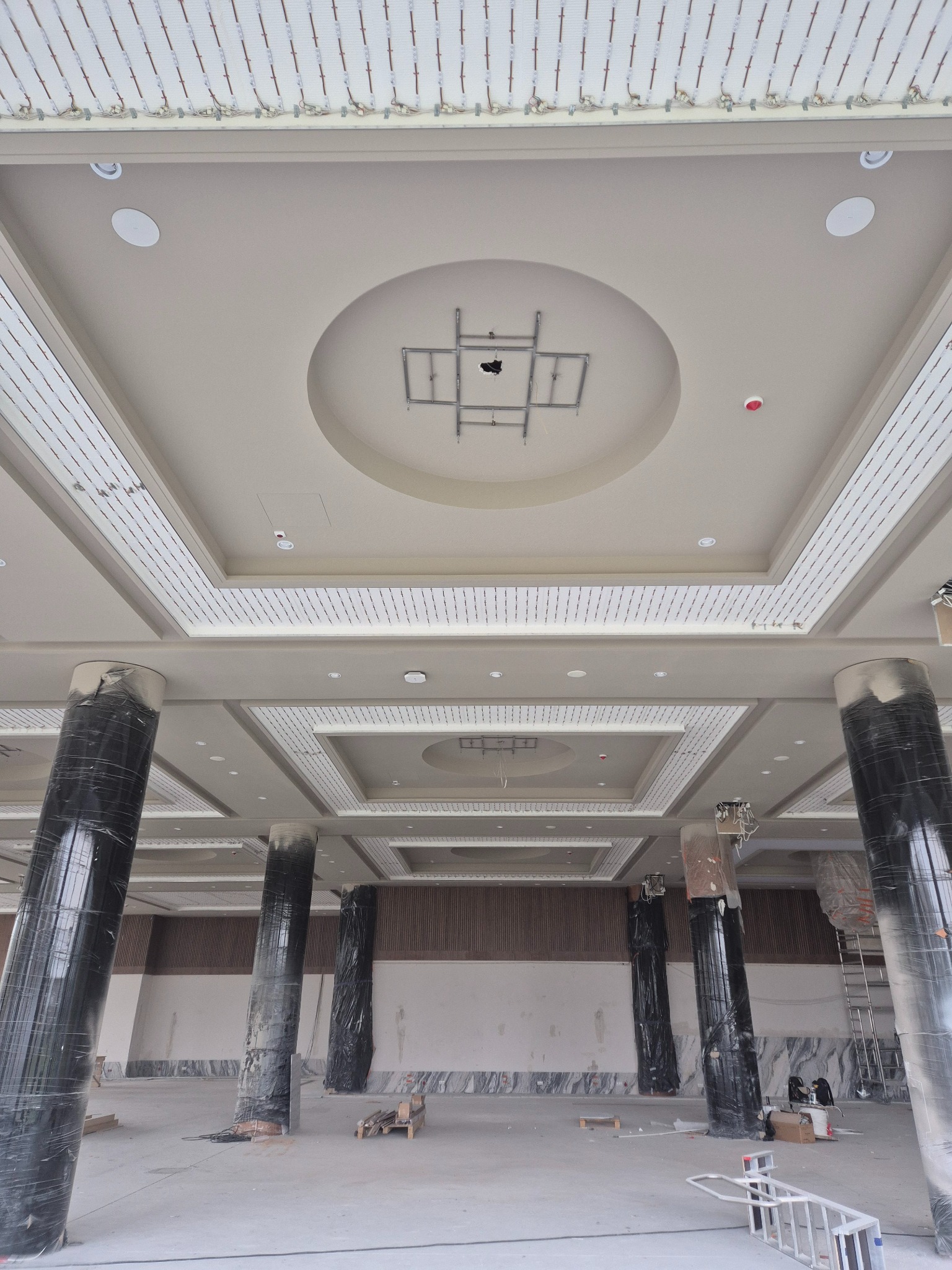 Wnętrze w trakcie wykończenia: dekoracyjny sufit z oświetleniem LED, kolumny zabezpieczone folią, drabina. Widok z dołu, szeroki kadr.