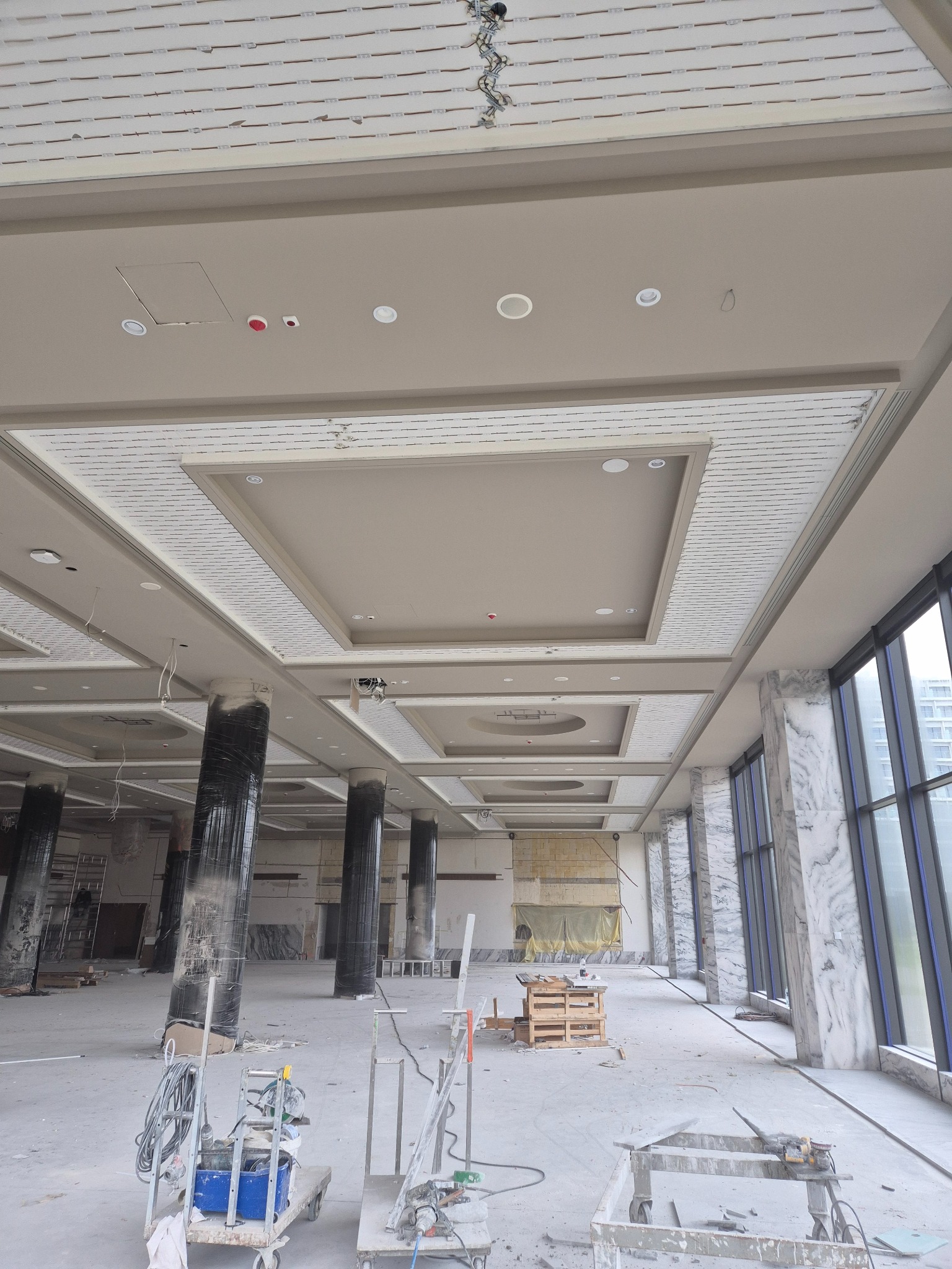 Wnętrze w trakcie montażu sufitu kasetonowego z oświetleniem punktowym. Widoczne narzędzia i materiały budowlane. Kolumny owinięte folią ochronną.