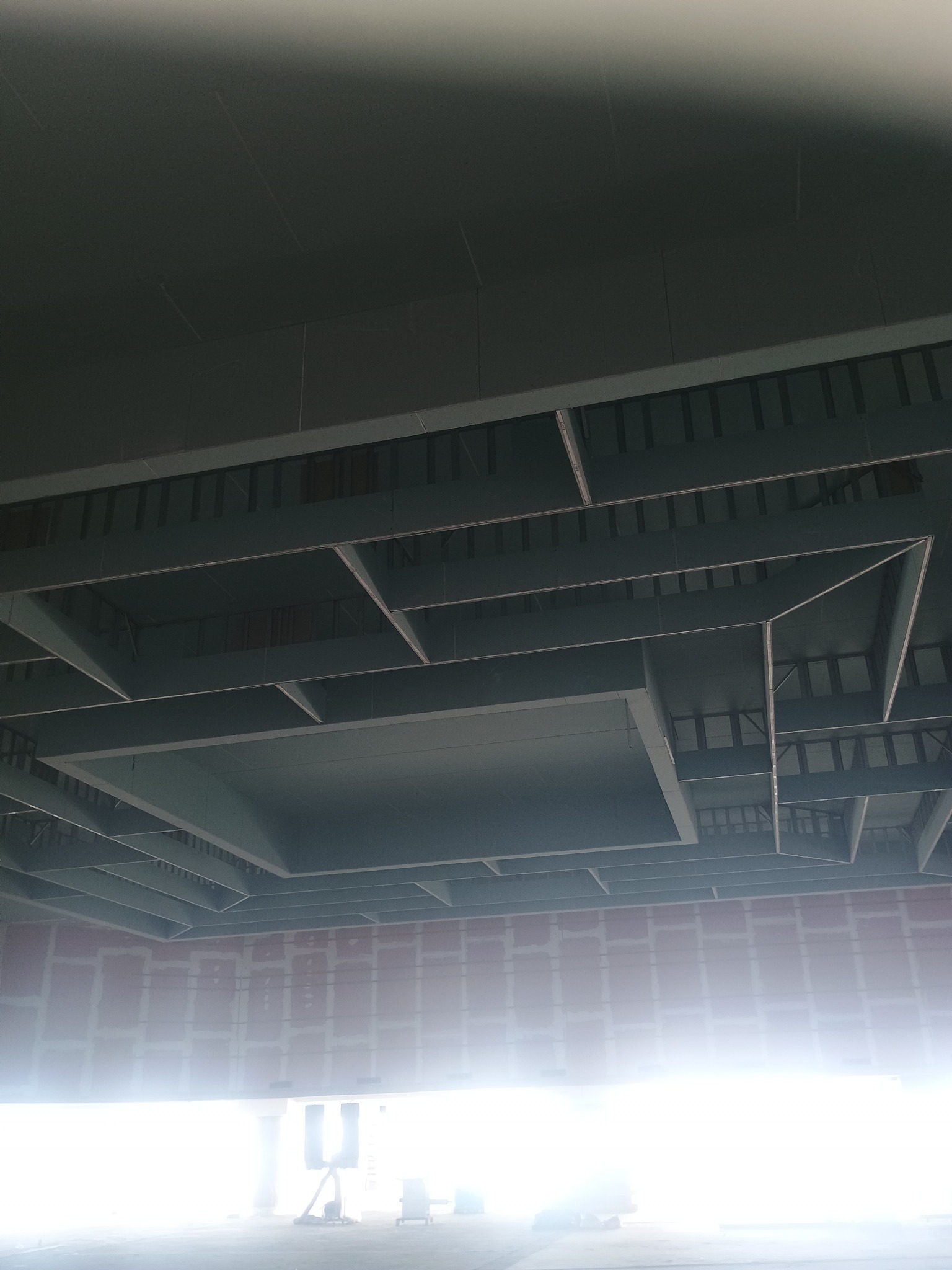 Sufit z widoczną konstrukcją nośną i częściowo zamontowanymi panelami. Widoczne łączenia płyt na ścianie. Oświetlenie od dołu.