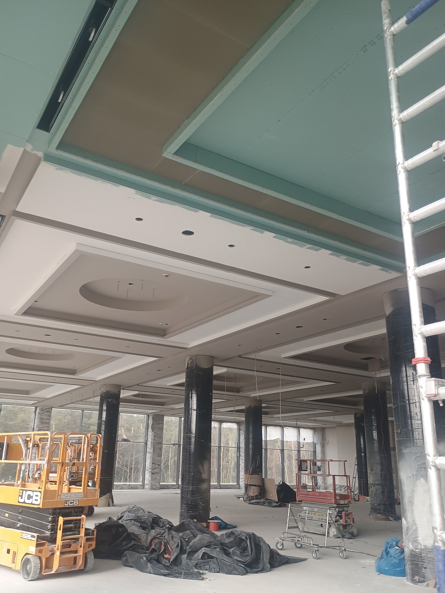 Montaż sufitu podwieszanego z płyt karton-gips w przestronnym wnętrzu. Widoczne elementy konstrukcyjne, kolumny i podnośniki. Etap prac wykończeniowych.