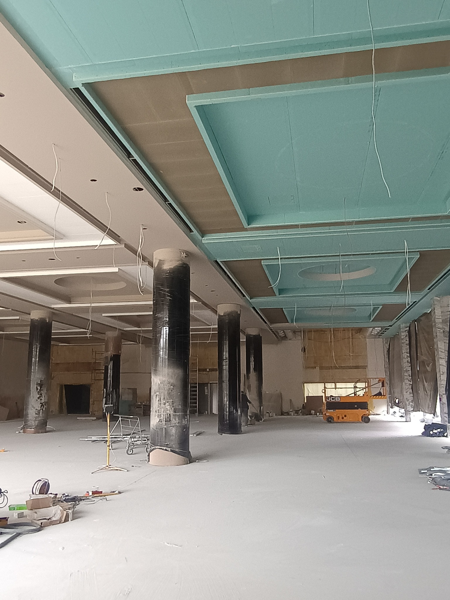 Montaż sufitu podwieszanego z płyt kartonowo-gipsowych w przestronnym wnętrzu. Widoczne kolumny i podnośnik. Etap prac wykończeniowych.