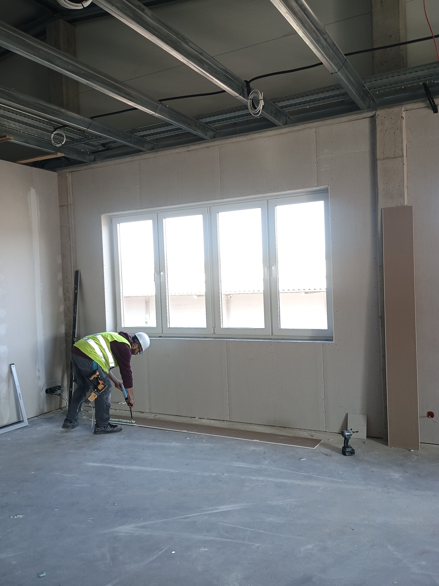 Montaż płyty gipsowej przy oknie: pracownik w kasku i kamizelce, narzędzia, surowy stan wykończenia, widoczne profile sufitowe. Wnętrze w trakcie prac.