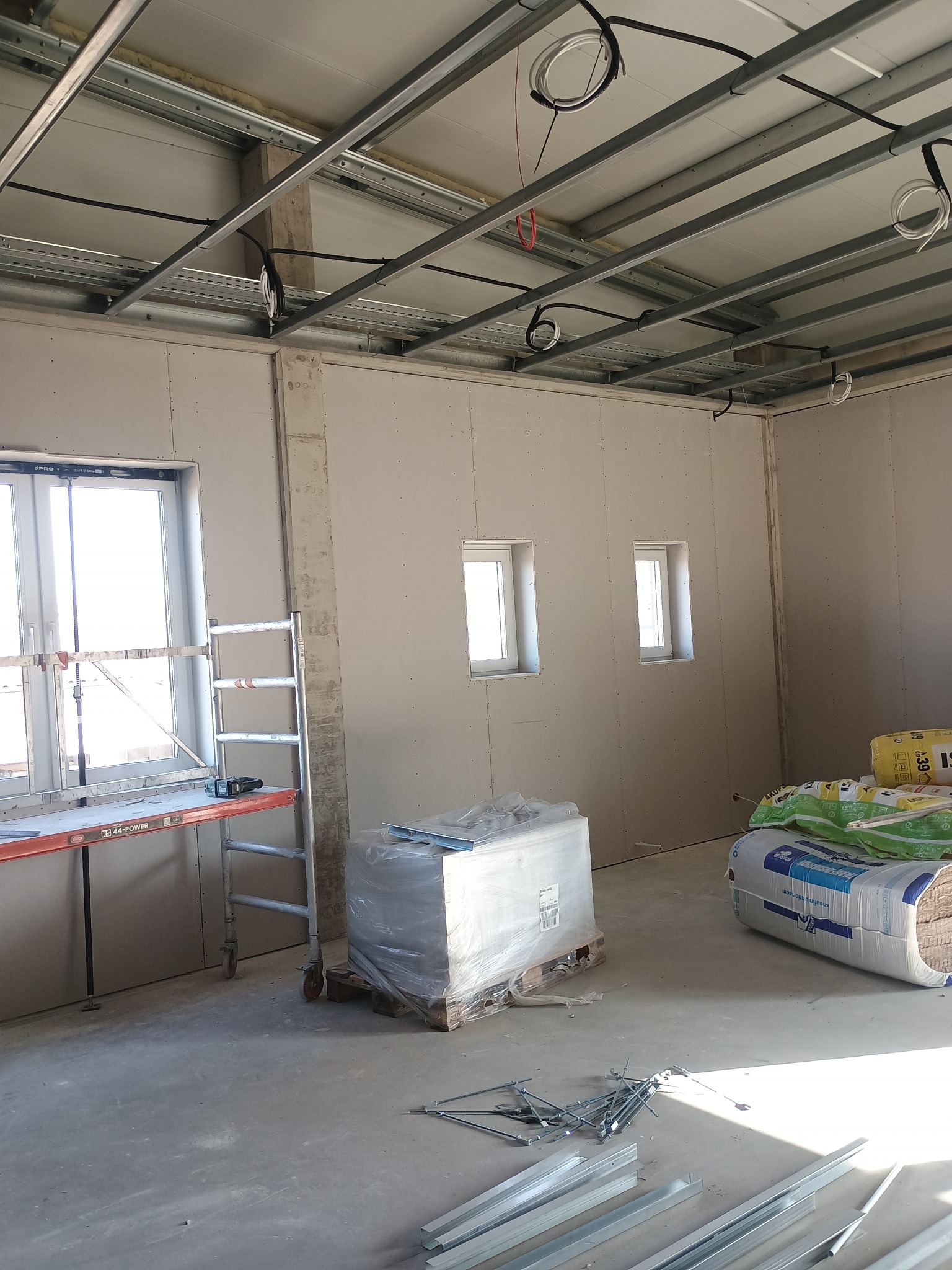 Montaż ścian z płyt gipsowo-kartonowych w nowym budynku. Widoczne profile, płyty, okna i materiały izolacyjne. Prace wykończeniowe w surowym wnętrzu.