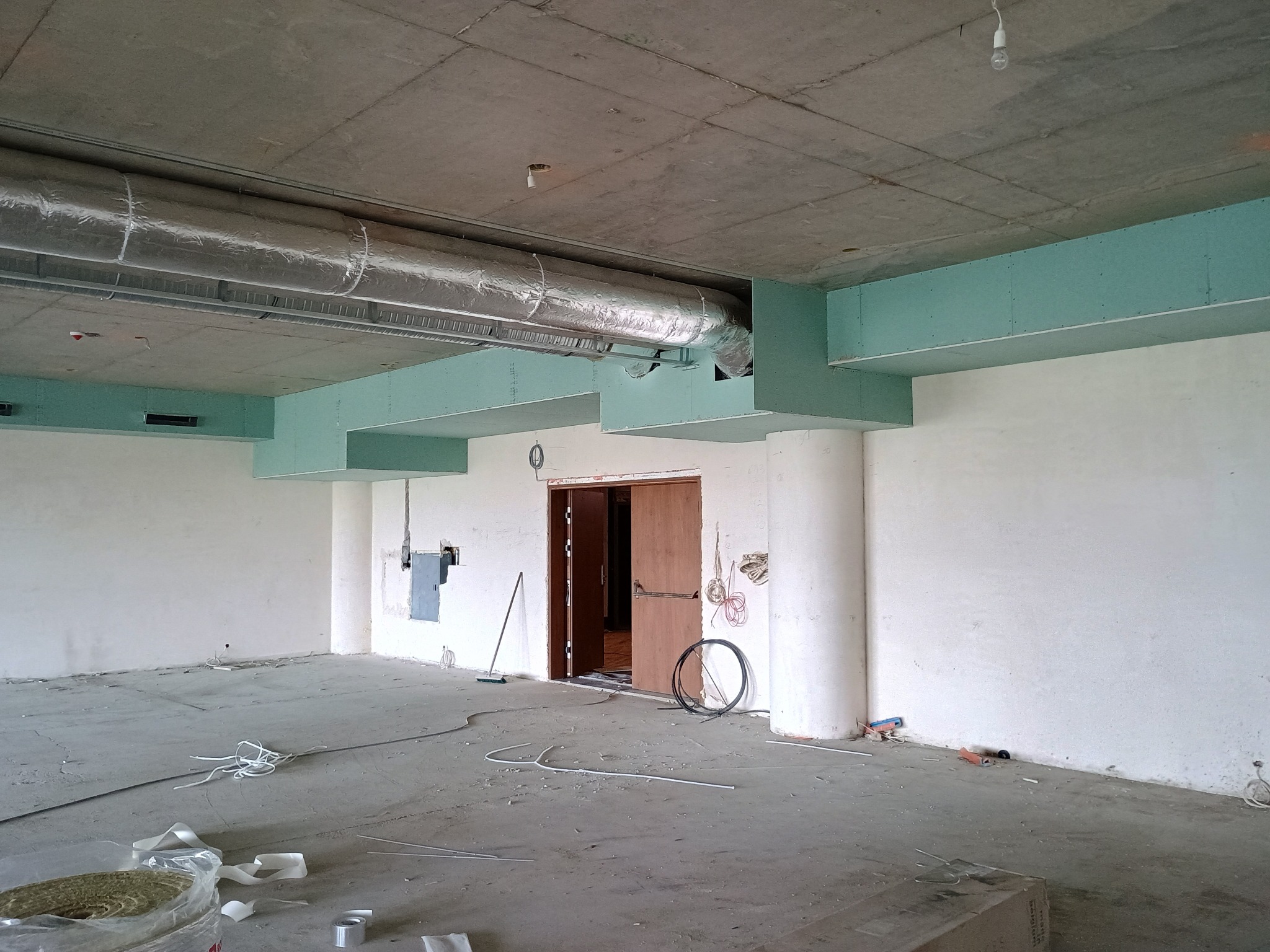 Wnętrze w trakcie prac wykończeniowych: surowy strop, instalacja wentylacyjna, fragmenty ścian z zielonymi płytami, przewody i materiały budowlane na podłodze.