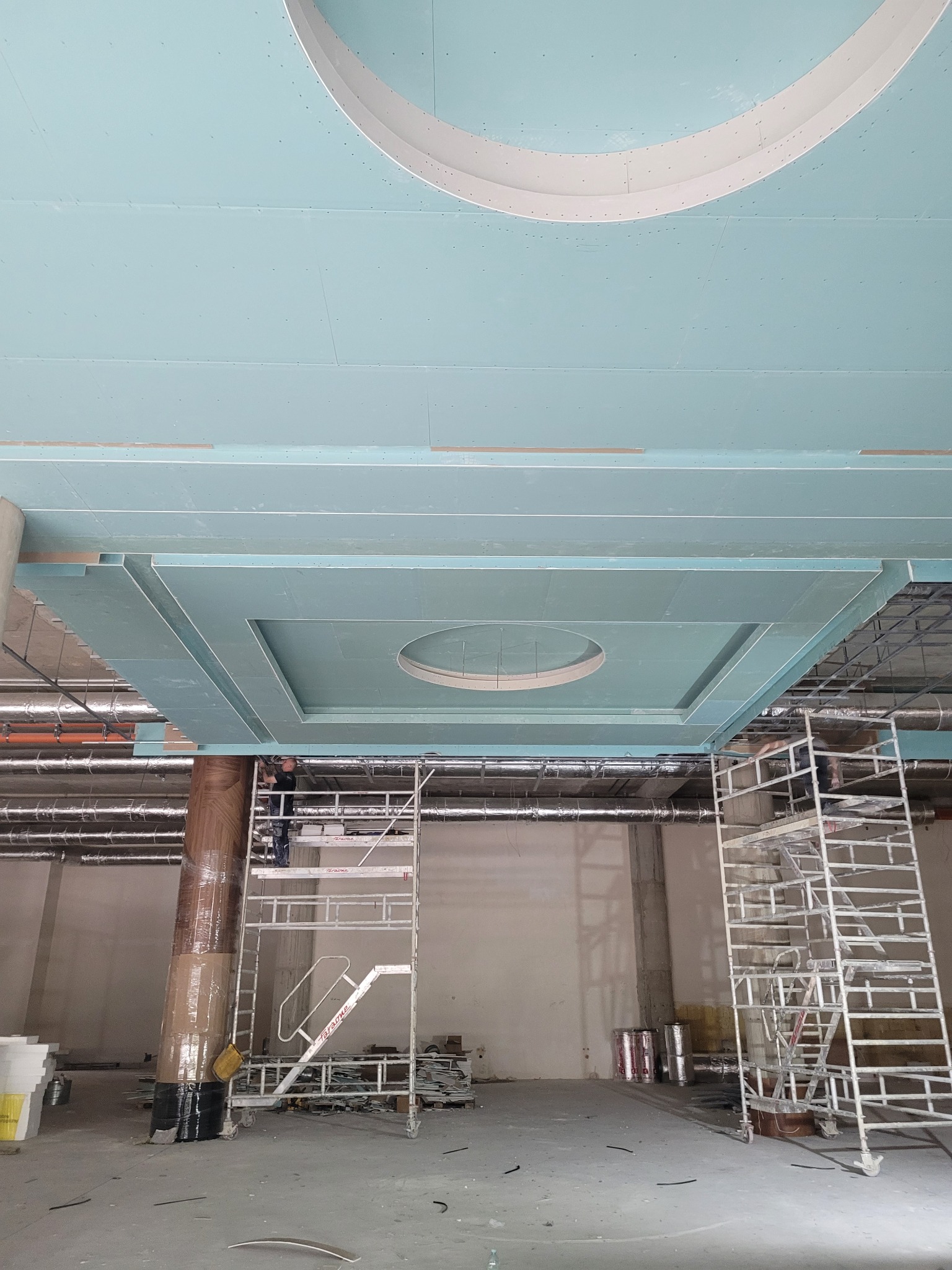 Montaż sufitu z płyty karton gips z elementami dekoracyjnymi w Koszalinie. Widoczne rusztowania i pracownik podczas pracy. Nietypowy kształt sufitu.