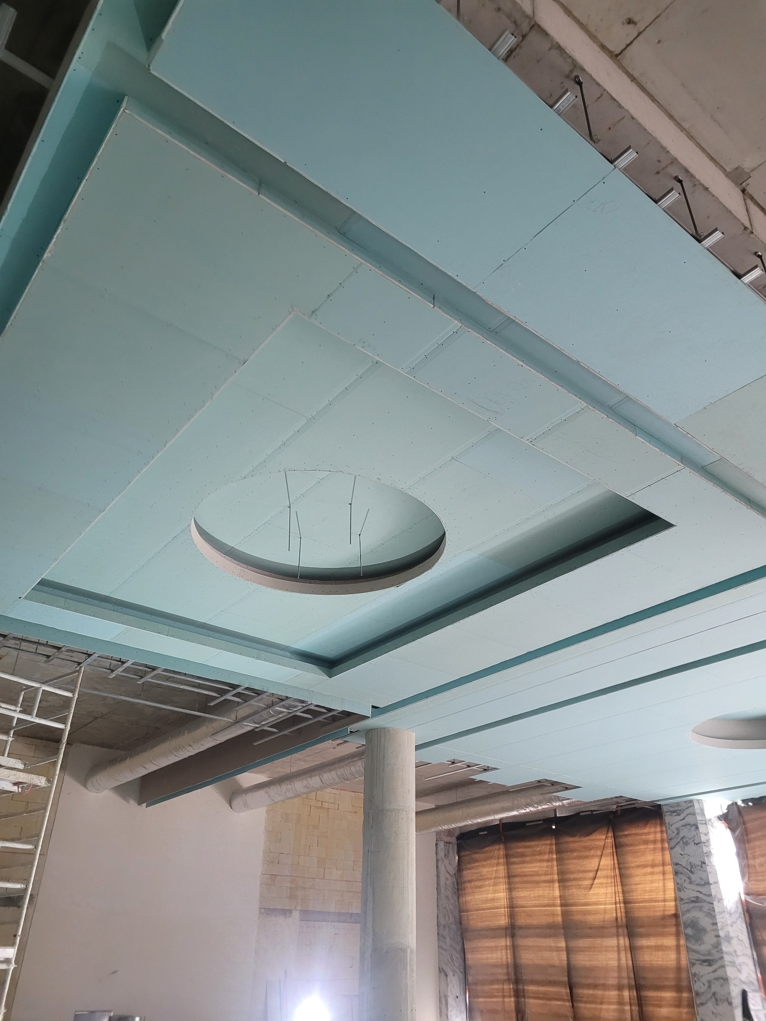 Niebieski sufit podwieszany z płyt kartonowo-gipsowych w trakcie montażu, widoczne profile i okrągłe wycięcie na oświetlenie. Częściowo widoczne rusztowania.