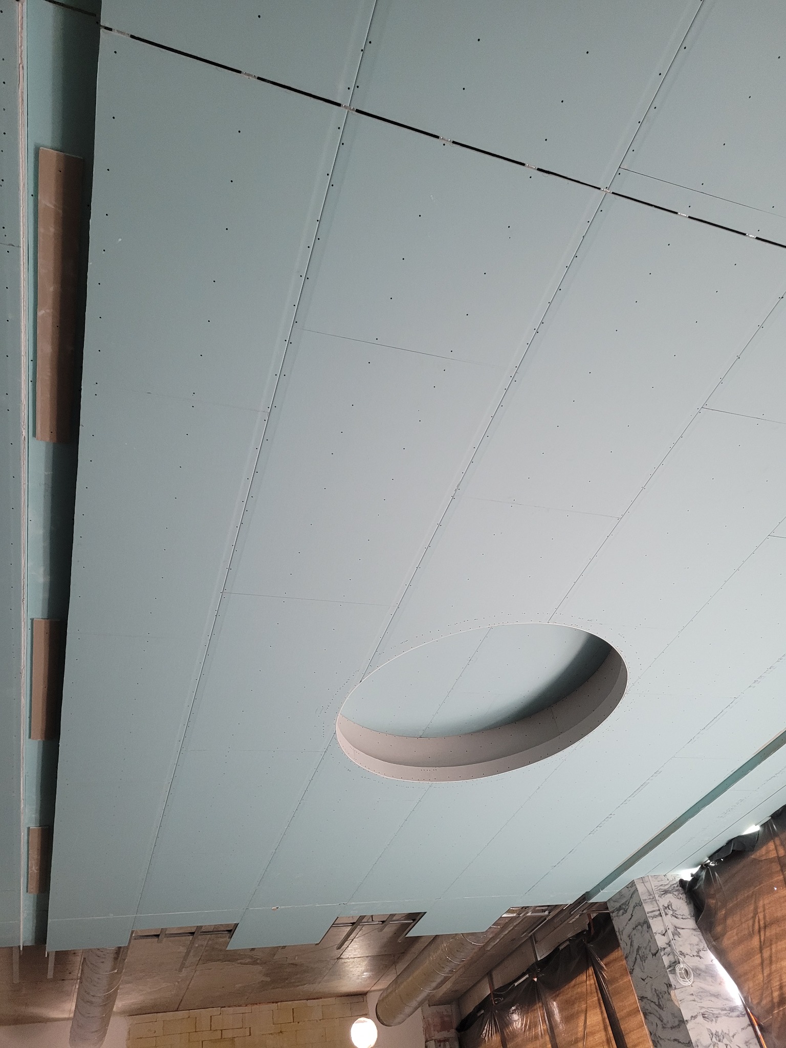 Montaż sufitu z płyt gipsowych z okrągłym otworem na oświetlenie. Widoczne łączenia płyt i konstrukcja nośna. Ujęcie z dołu, perspektywa zbieżna.