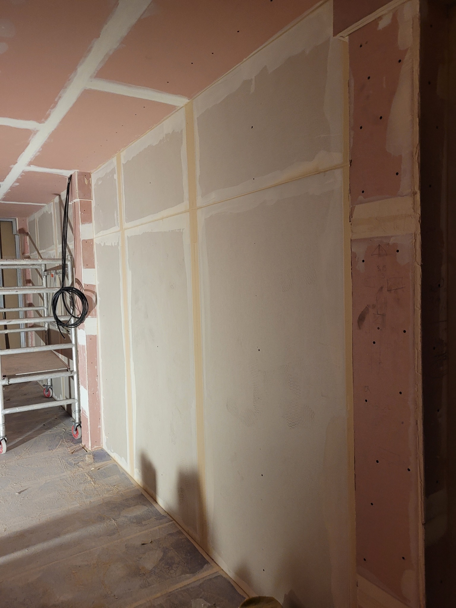 Wnętrze w trakcie prac wykończeniowych: ściany z płyt gipsowo-kartonowych, widoczne profile i szpachlowanie. W tle rusztowanie i przewody elektryczne.
