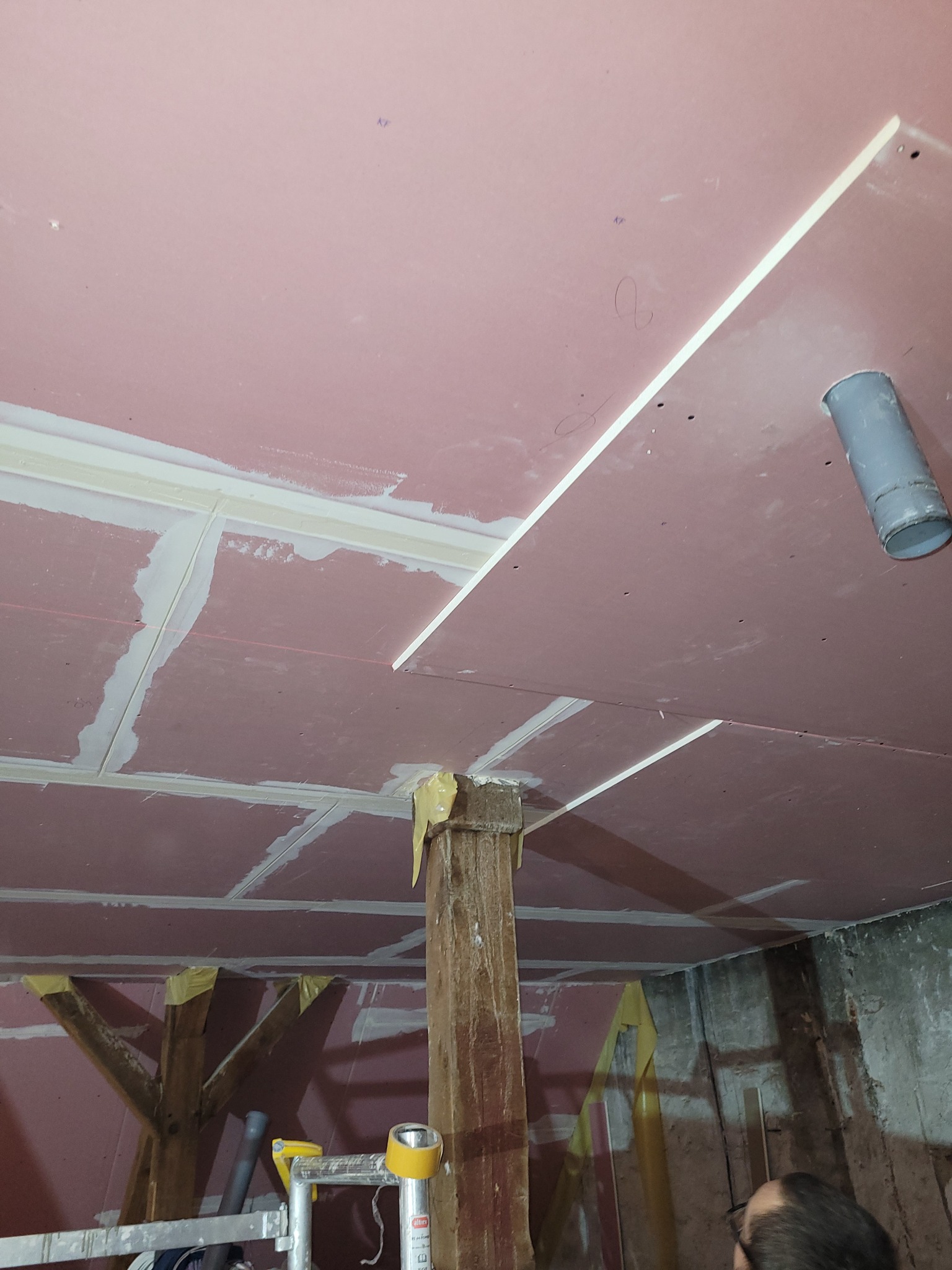 Montaż płyt na suficie podwieszanym z widocznymi elementami konstrukcyjnymi i rurą wentylacyjną. Prace wykończeniowe w trakcie realizacji.