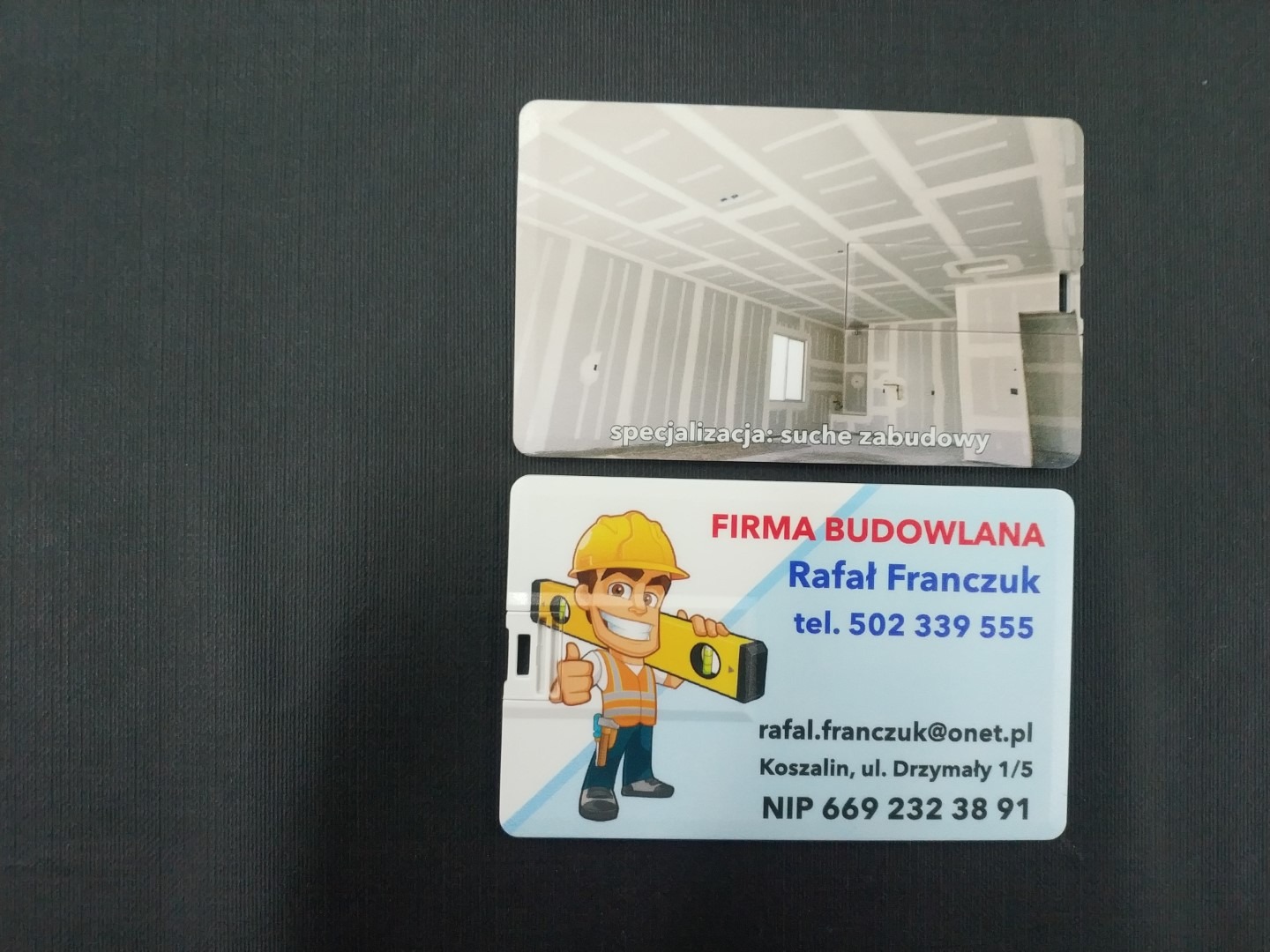 Wizytówka firmy budowlanej z ilustracją wnętrza w trakcie montażu suchej zabudowy i danymi kontaktowymi, na ciemnym tle.