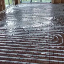 Instalacja ogrzewania podłogowego z rur na folii z siatką w surowym drewnianym budynku w trakcie budowy, widok przez okno na zewnątrz.