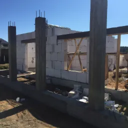 Wznoszenie ścian z betonu komórkowego na fundamencie z betonowymi słupami nośnymi w trakcie budowy domu, widoczne zbrojenia pionowe i podkłady pod okna.