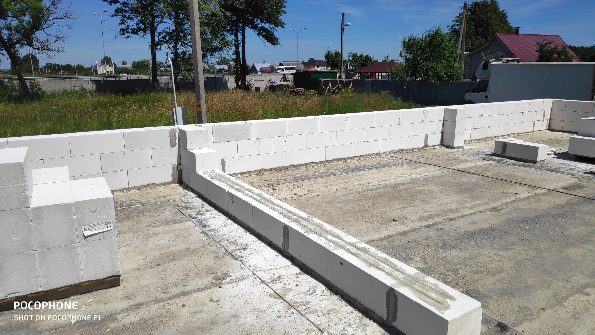 Wznoszenie ścian fundamentowych z białych bloczków betonowych na wylewce, widoczne zbrojenie w spoinach, w tle krajobraz z drzewami i drogą.