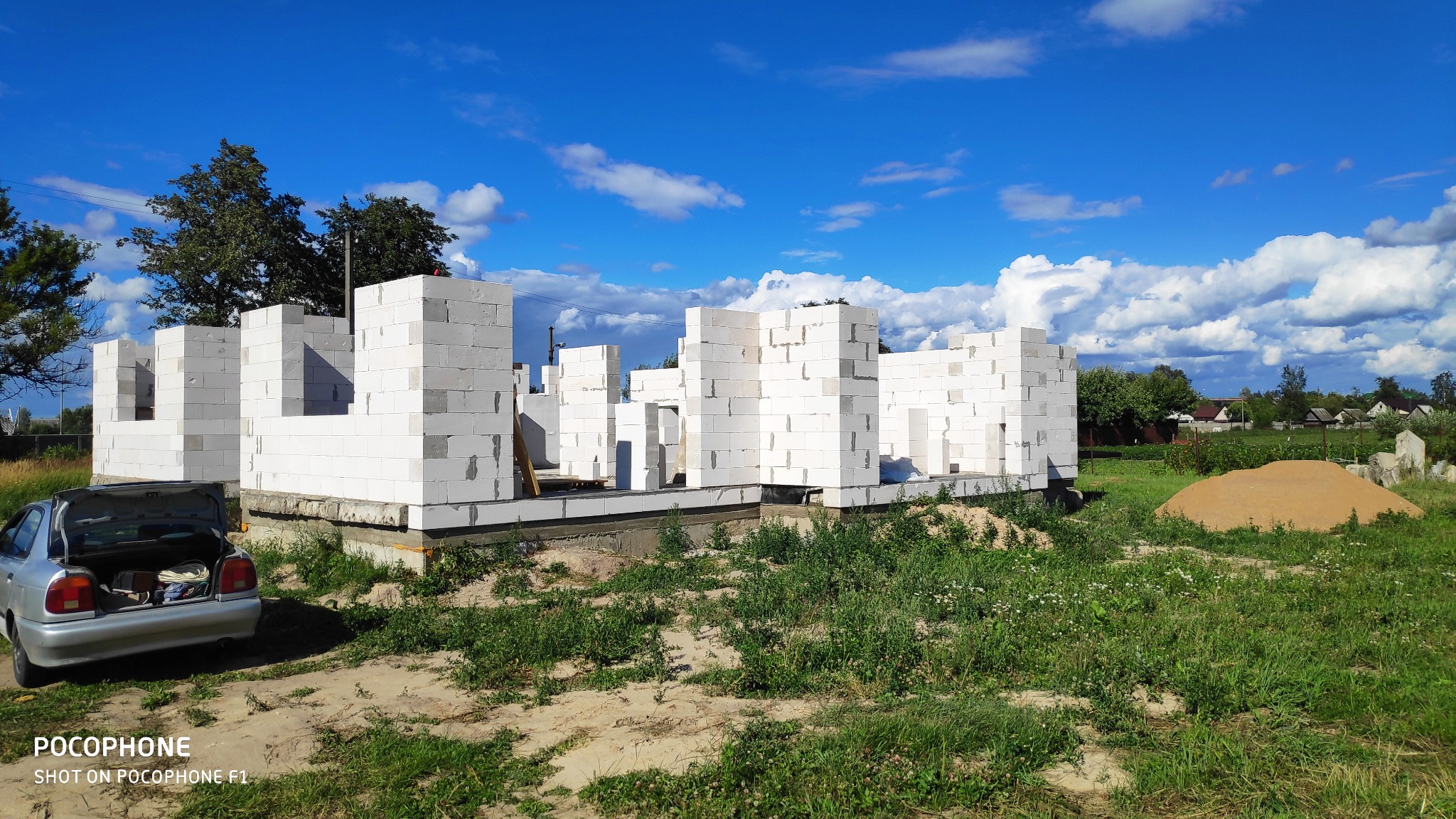 Wznoszone ściany parteru z białych bloczków betonu komórkowego, widoczny fundament, otwarty bagażnik samochodu osobowego i piasek na działce budowlanej, błękitne niebo z obłokami w tle.