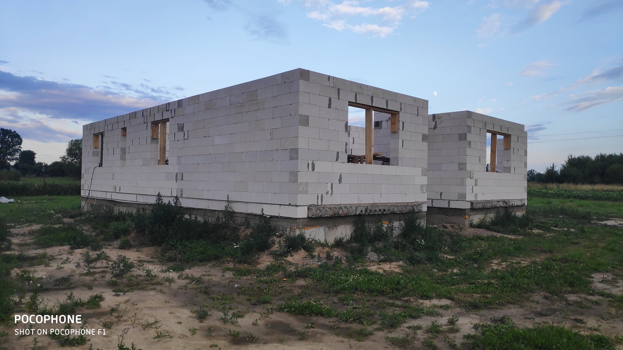 Wznoszenie ścian parterowego domu z białych bloczków, widoczne drewniane ramy okienne, teren zarośnięty trawą, pochmurne niebo w tle.