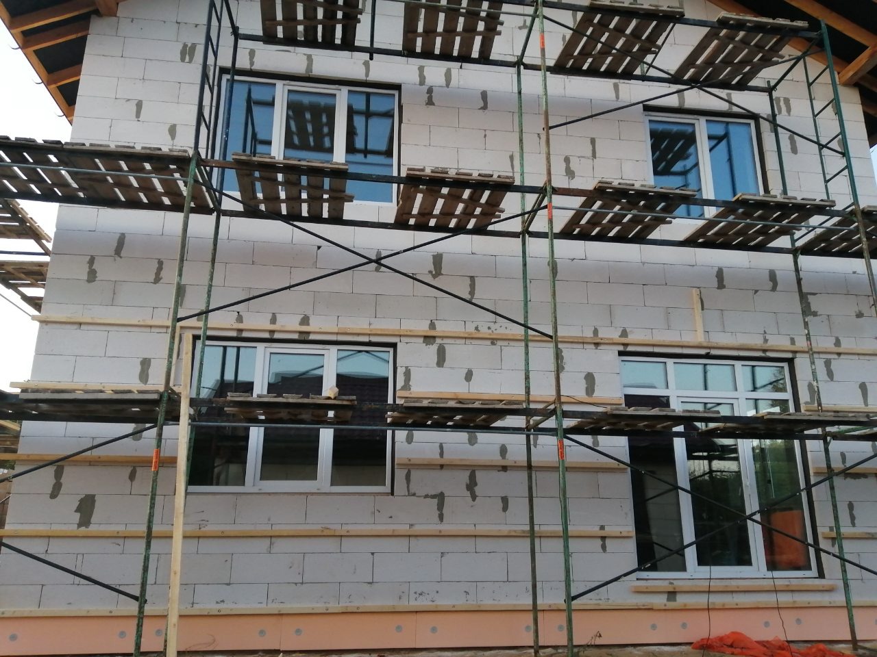 Budowa domu z białych bloczków, widoczne rusztowanie i wstawione okna, dach w trakcie budowy.