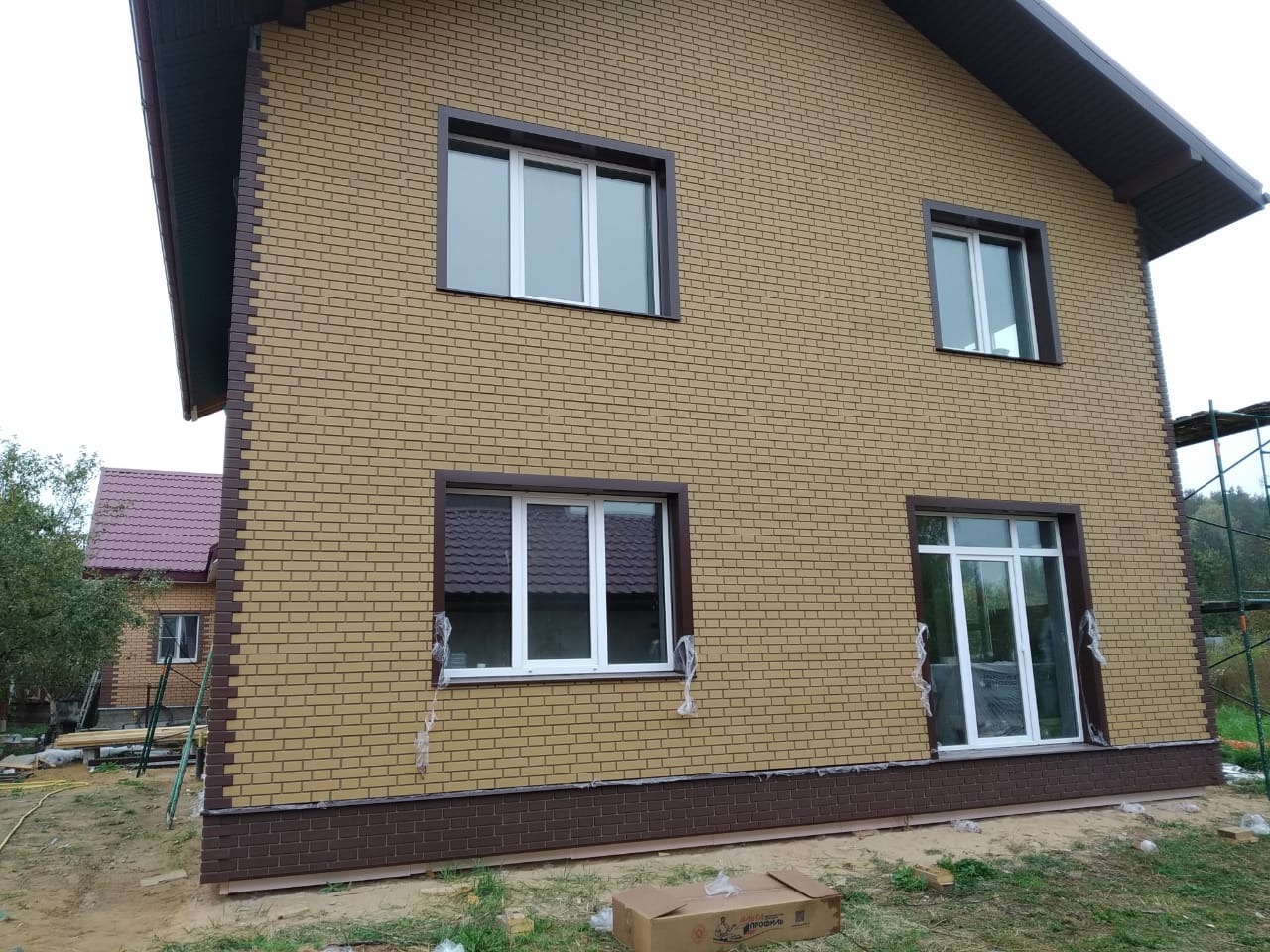 Dwukondygnacyjny dom z elewacją imitującą cegłę w odcieniach beżu i brązu, z białymi oknami i częściowo zdjętą folią ochronną, widoczny karton na trawie i fragment rusztowania w tle.