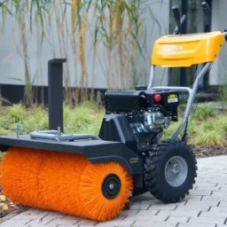 Żółto-czarna zamiatarka spalinowa Stiga z pomarańczową szczotką na kostce brukowej, w tle trawa ozdobna i nowoczesna architektura.