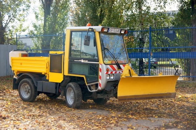 Żółty pojazd do odśnieżania z pługiem, zaparkowany na liściach w Krakowie, na tle niebieskiego ogrodzenia i drzew.
