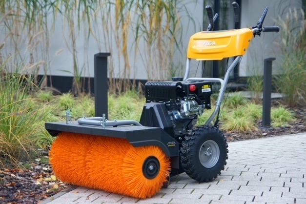 Żółto-czarna zamiatarka spalinowa Stiga z pomarańczową szczotką na kostce brukowej, w tle trawa ozdobna i nowoczesna architektura.