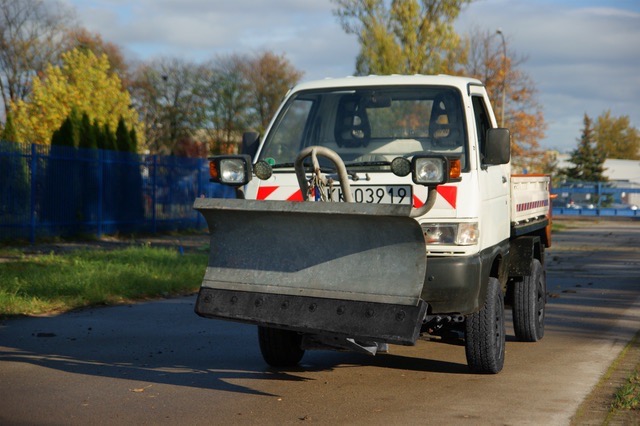 Mała biała ciężarówka z zamontowanym pługiem śnieżnym, zaparkowana na asfaltowej drodze w jesienny dzień, widoczne drzewa w tle.